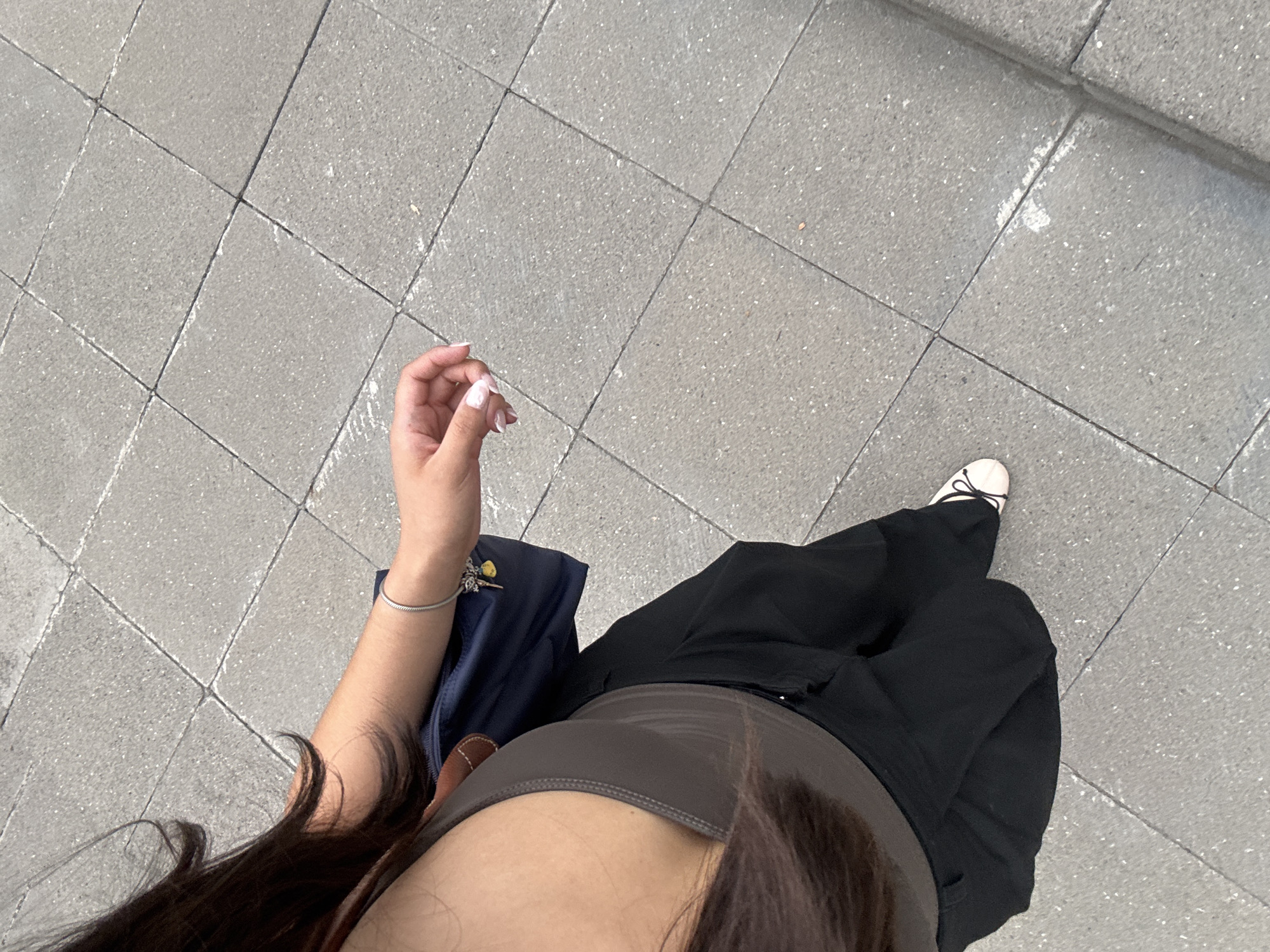 Here's a description of the image:
**Scene**: A first-person perspective photograph looking down. The ground is paved with grey square concrete tiles arranged in a grid pattern.
**Subject**: The subject is wearing black pants and a brown top. A portion of a dark blue bag is visible near the subject's left side. The subject's left hand is in view, and a silver bracelet is around the wrist. A single white shoe is visible on the right side. Long dark brown hair frames part of the view.
**Location**: Without additional clues in the photograph, it is not possible to confirm that the location is Puebla, Mexico.
