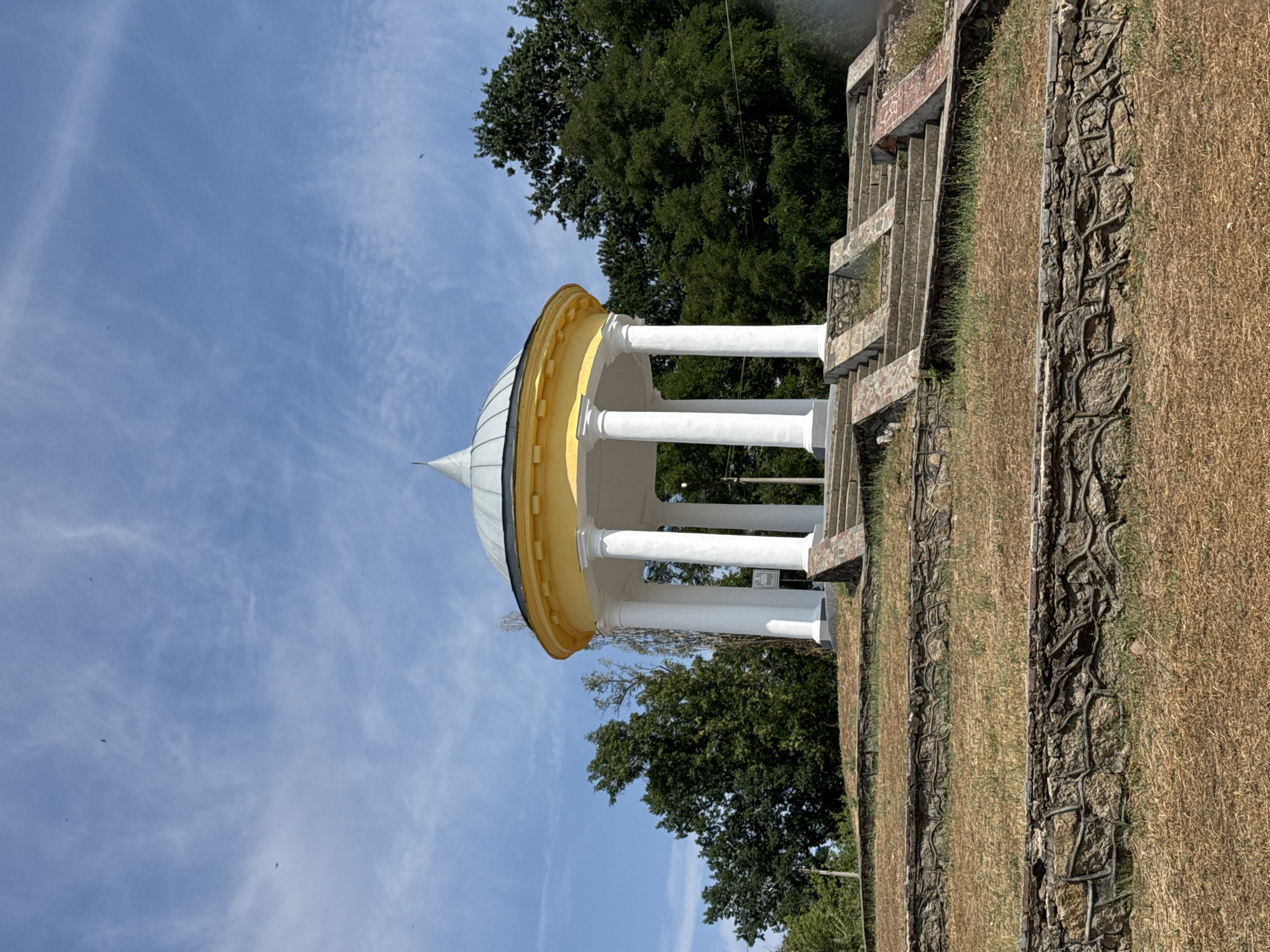 A gazebo with a yellow roof and white columns sits atop a tiered, grassy hill. The gazebo is surrounded by a cluster of tall, green trees. Stone retaining walls and concrete steps lead up to the gazebo. The sky above is blue with wispy white clouds, and several small dark shapes, likely birds, are visible in the sky. The scene appears to be in a park or public space. This is likely the Voznesensk ArboRetum in Voznesensk, Ukraine, a public park known for its collection of trees and the gazebo, which is a popular landmark.
