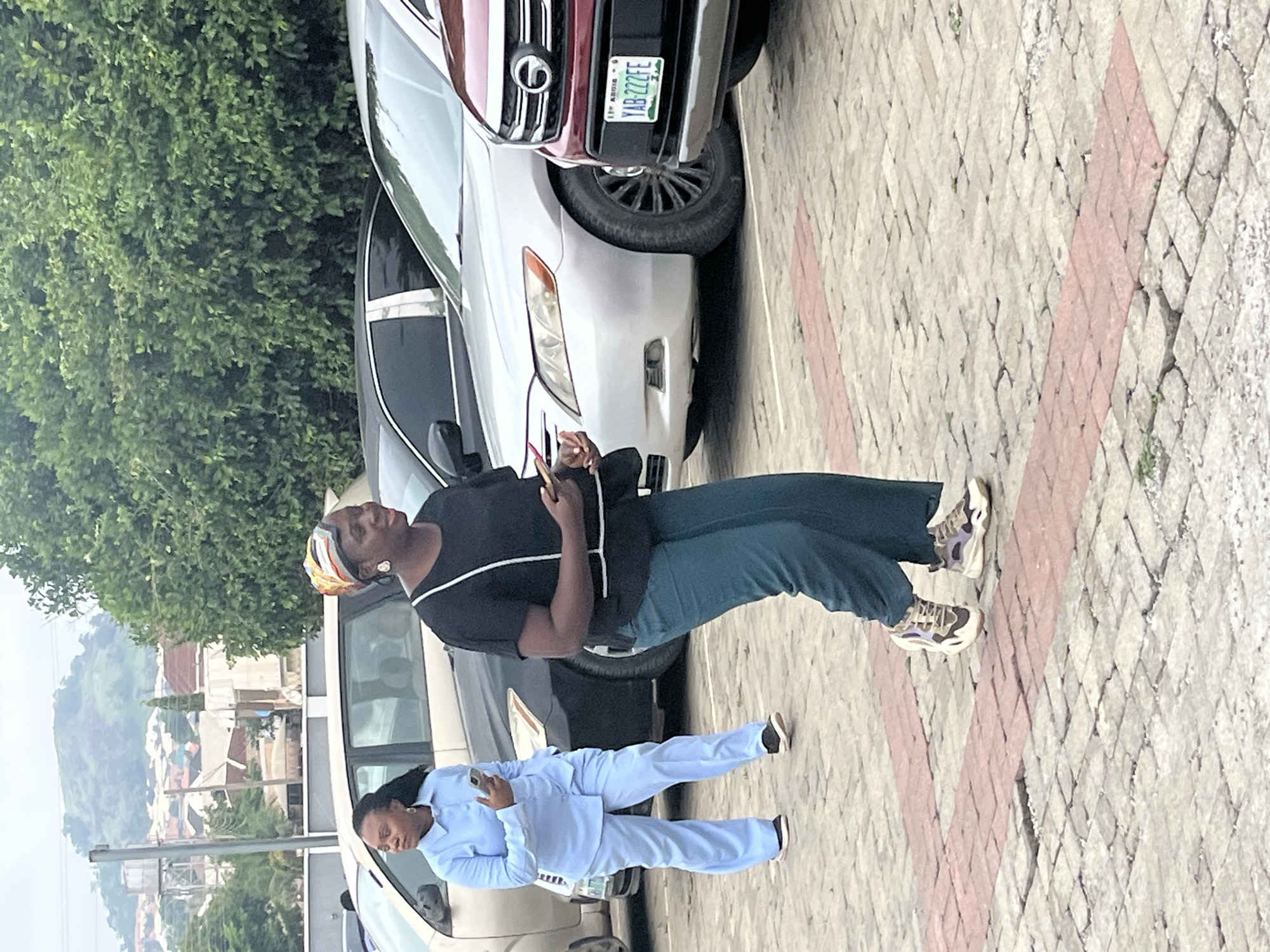 In the city of Abuja, Nigeria, on a cloudy day, two women are in a parking lot. The woman on the left is wearing a light blue pantsuit and holding a smartphone. The woman on the right is wearing a black top with white trim and teal pants, and is also holding a smartphone. Several cars are parked in the lot, including a silver sedan and a red SUV with a Nigerian license plate. The background shows trees and buildings, with a hill in the distance. The ground is paved with interlocking bricks.