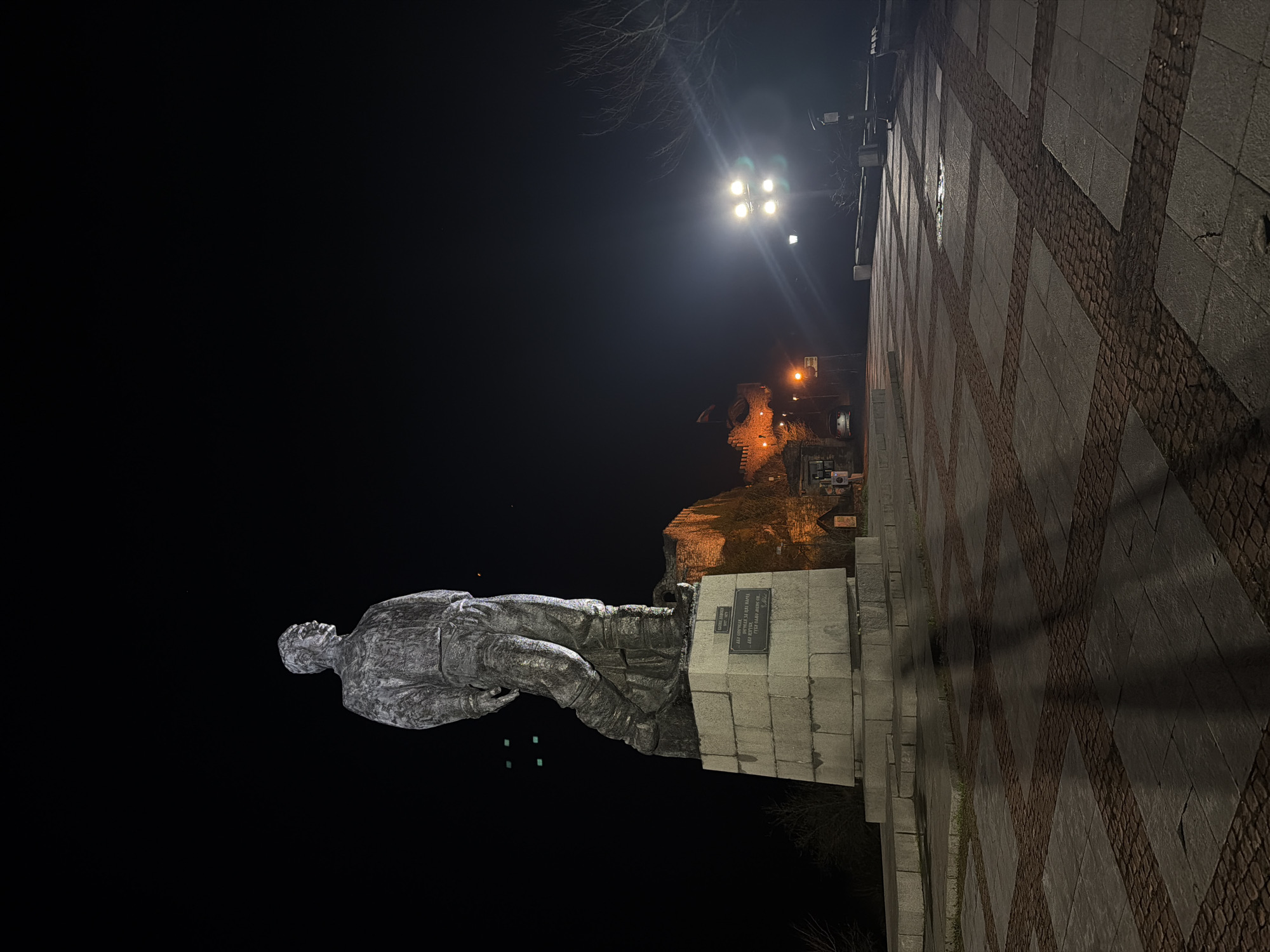 A nighttime shot depicts a bronze statue of Vasil Levski, a Bulgarian revolutionary hero, standing atop a stone pedestal. The monument is located in Lovech, Bulgaria, in front of the Lovech Fortress. The foreground consists of a patterned stone pavement leading up to the monument. Above the monument is a modern lighting fixture, and behind it, the Lovech Fortress is illuminated. The night sky is dark.