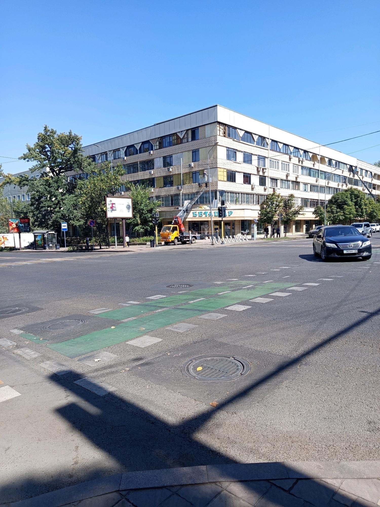This is a wide-angle, eye-level shot of a street scene in Almaty, Kazakhstan, on a clear, sunny day. The foreground is dominated by a multi-lane intersection with a marked crosswalk, distinguished by green painted lanes and white squares. A circular manhole cover is visible in the asphalt. In the midground, to the right, a dark-colored sedan, possibly a Toyota Camry, with license plate "382 BHT 02", is turning at the intersection. Across the street, a large, light-colored, multi-story building with numerous windows and air conditioning units lines the horizon. In front of this building, a yellow flatbed truck with a crane is parked, with its boom extended upwards. There are a few pedestrians visible near the building and a bus stop on the left side of the street. Trees are scattered throughout the scene, with a particularly large one on the left. The overall impression is of an urban environment on a bright, busy day.