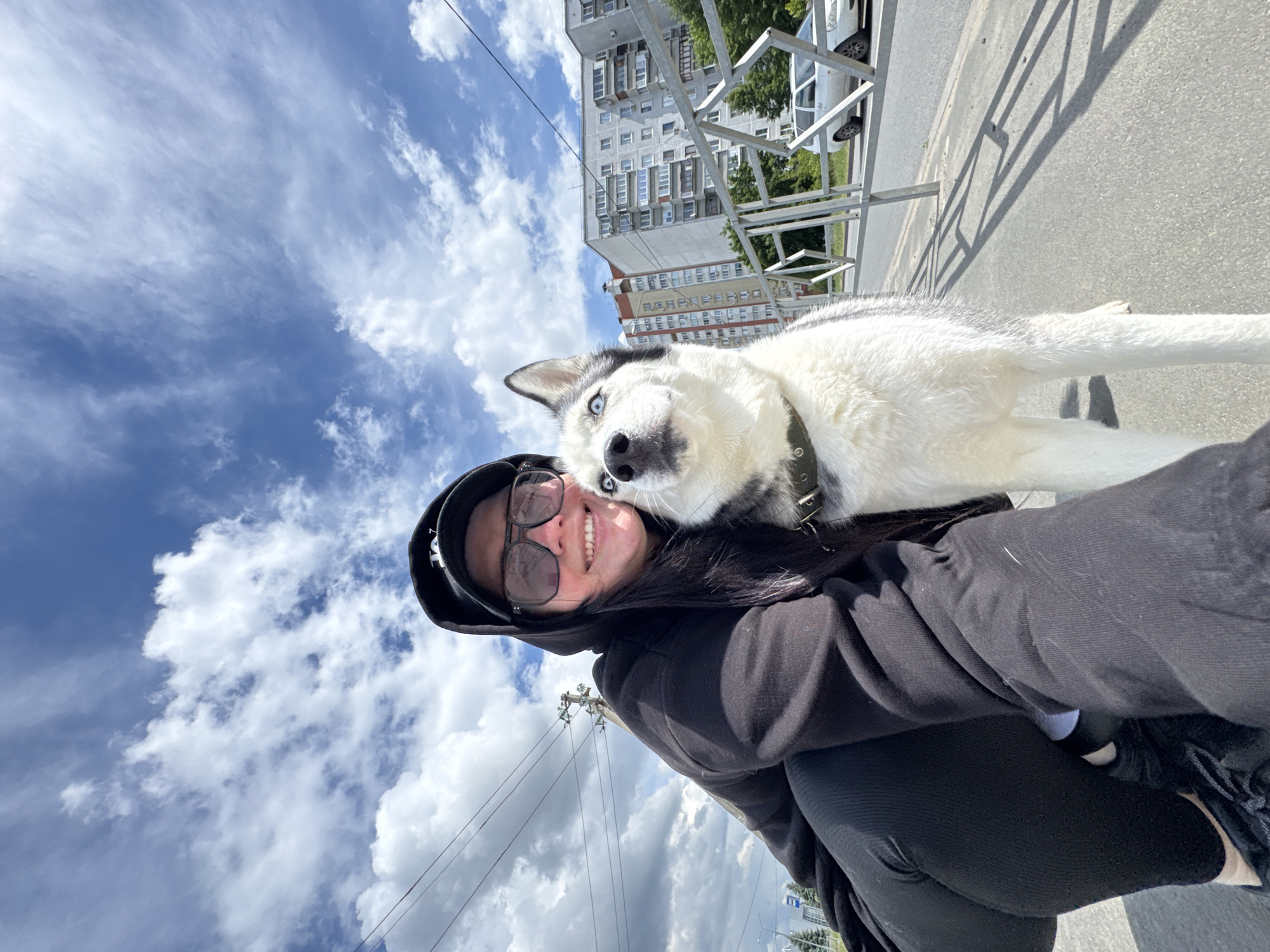 A medium shot captures a fair-skinned individual with long, dark hair, dark glasses, and a black hoodie, positioned against a backdrop of a bright blue, cloudy sky. The individual smiles with teeth showing. A husky dog with piercing blue eyes rests its head on the individual’s shoulder.
The scene also features a light-colored building with multiple windows in the background and a metal structure with vertical bars. A light-colored vehicle is partially visible behind the metal structure. The scene is oriented at an angle. The location appears to be an urban environment. Power lines are visible.
