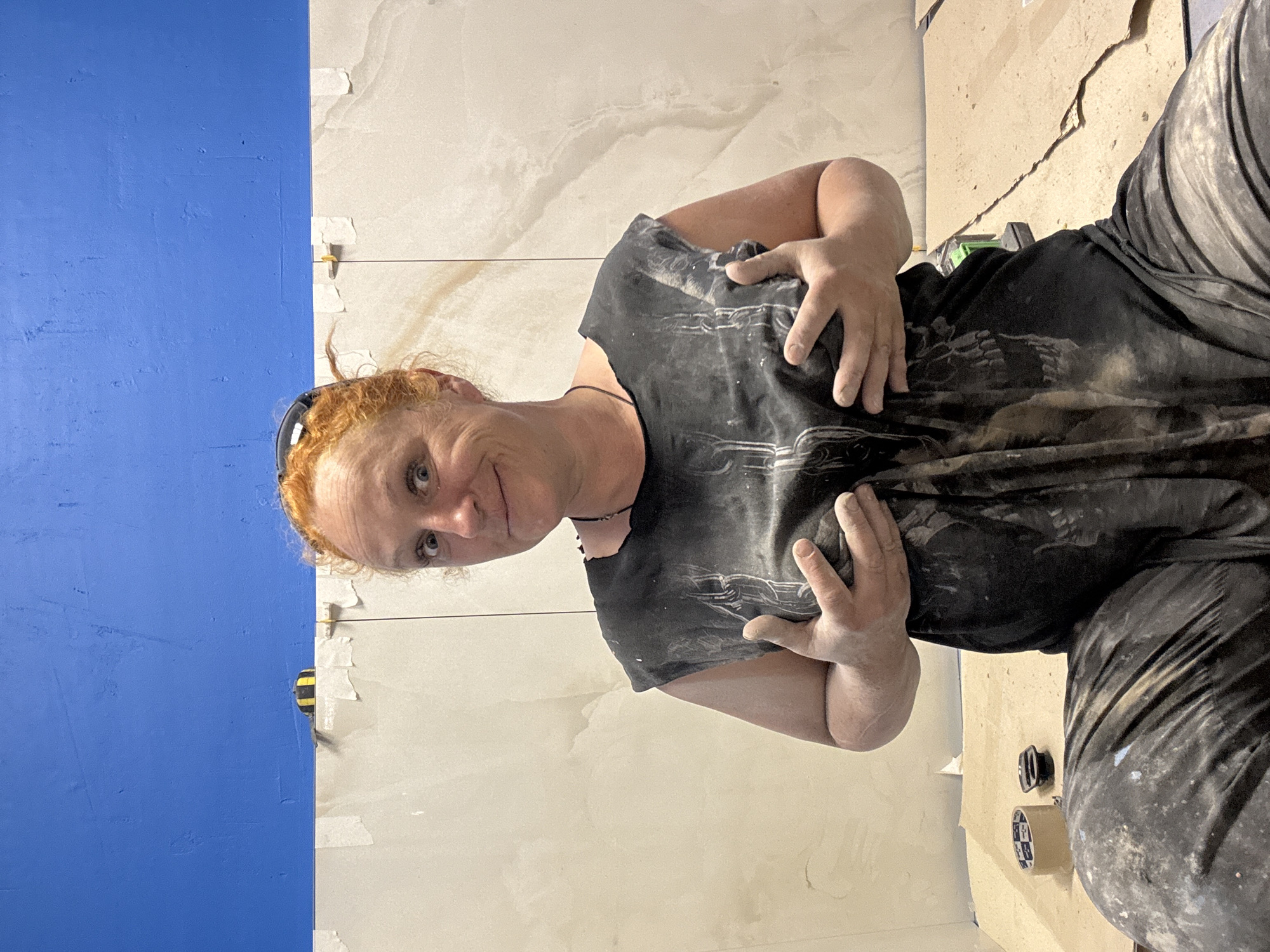 A woman with red hair and blue eyes stands in front of a wall partially tiled with large, light-colored tiles featuring a marbled pattern. A section of the wall on the left is painted bright blue. The woman is wearing a dark-colored T-shirt and dark pants, both covered in what appears to be dust or plaster. She is looking at the camera with a slight smile and her hands are positioned in front of her torso. On the floor, various items are visible, including tools, tape, and tile spacers, indicating that she is likely in the process of tiling the wall. The location of the scene is not discernable from the image, and there is no verifiable information indicating it is located in Chaikovsky, Russia.