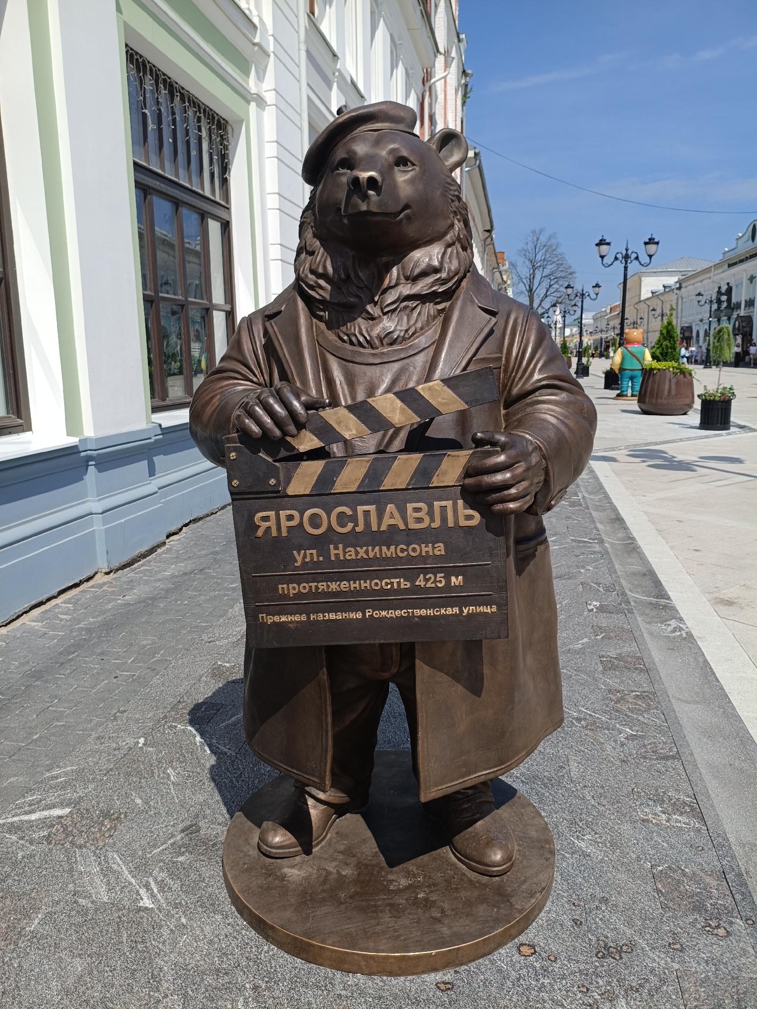 A bronze statue of a bear wearing a beret and a trench coat stands on a cobblestone street in Yaroslavl, Russia, holding a clapboard. The clapboard has text in Russian that reads "YAROSLAVL", "ul. Nakhimsona", "length 425 m", and "former name Rozhdestvenskaya street". The statue is in front of a building with large windows. In the background, there are people walking on the street, lampposts, and trees. The sky is blue and clear.