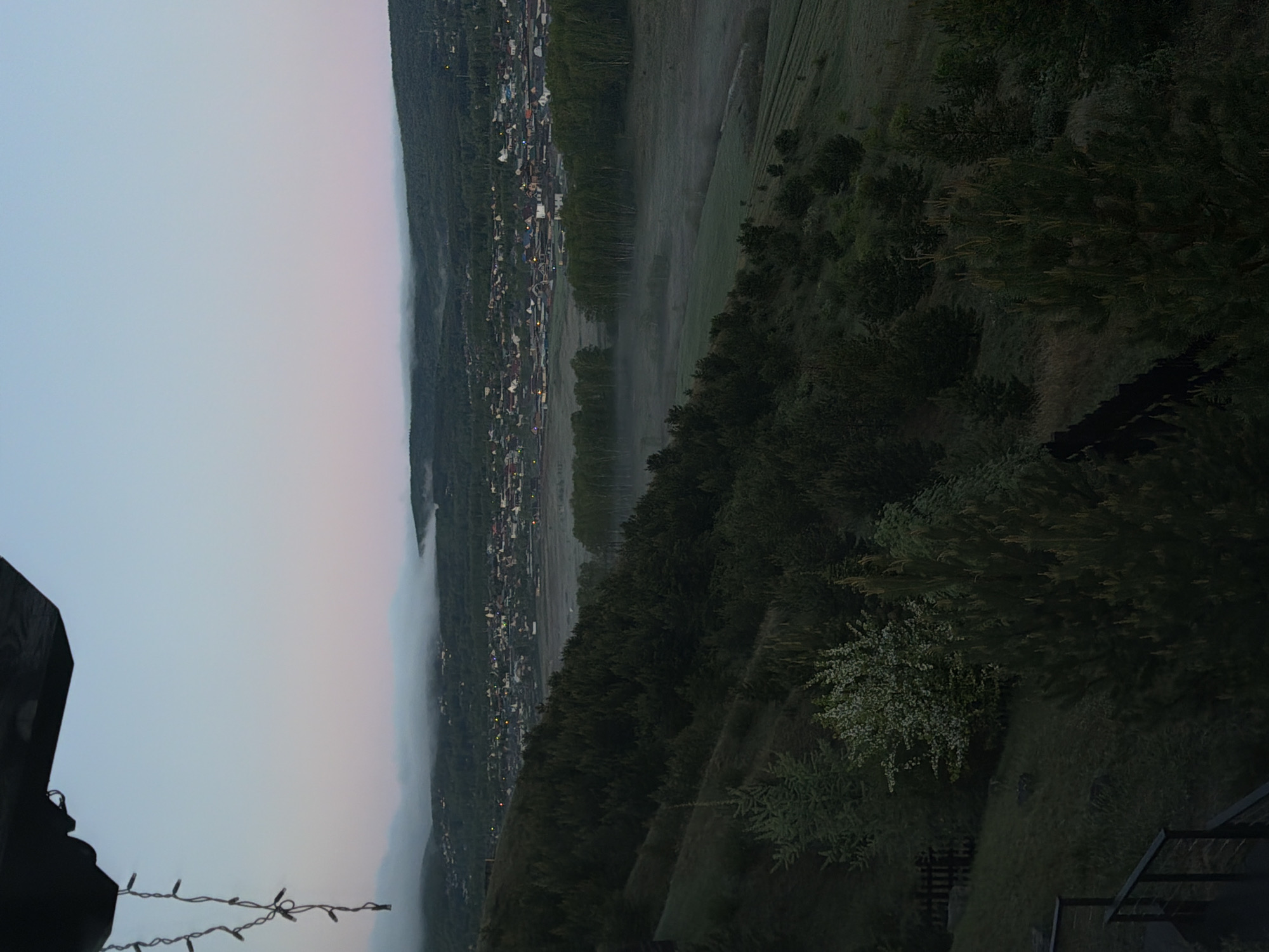 The image shows a view from a high vantage point, likely a hillside or elevated structure, looking down on a town in the valley in the early morning or late evening. A layer of fog partially obscures the view and hangs over the town, through which streetlights are visible. The landscape is characterized by trees on the right side of the image. On the left side of the image, the sky is visible. A string of lights is hanging on the bottom left of the image. The image is taken outdoors and the location is Shamoni, Russia.
