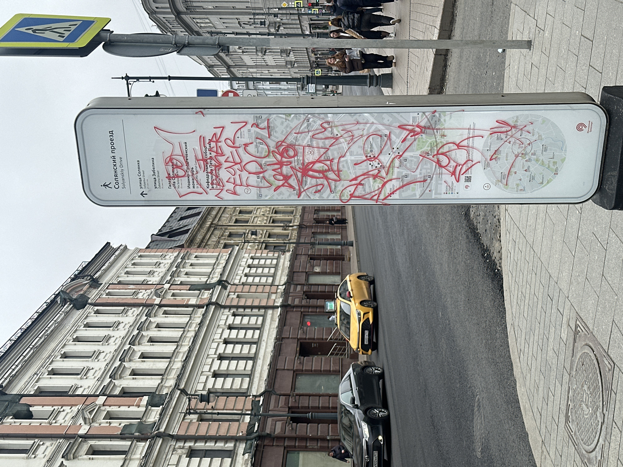 A daytime street scene in Moscow, Russia, captured on an overcast day, features a prominent tall, rectangular information stand heavily defaced with vibrant red graffiti. The stand displays a city map and tourist information in both Russian and English, identifying locations such as "Солянский проезд" (Solyanskiy Drive), "улица Солянка" (Solyanka Street), "улица Забелина" (Zabelina Street), "Галерея «На Солянке»" (Na Solyanke Art Gallery), "Иоанно-Предтеченский монастырь" (St John the Baptist Convent), and "Кафедральный собор апостолов Петра и Павла" (Cathedral of Saints Peter and Paul). The "Московский транспорт" (Moscow Transport) logo is also visible.
In the background, several multi-story buildings with classic, ornate architectural details line the street. One building on the left features a light-colored facade with numerous windows and decorative elements, while a darker, possibly brick, building stands behind the vehicles. Pedestrians can be seen walking on the sidewalk in the mid-ground.
On the asphalt road, a yellow taxi, likely a Yandex.Taxi given its visible branding, is parked or driving, followed closely by a black car. Part of the black car's license plate, "922A72", is discernible. A blue and yellow pedestrian crossing sign is mounted on a pole above the information stand. The ground consists of paving stones on the sidewalk and asphalt on the road, with visible manhole covers. The overall atmosphere suggests a typical, busy urban environment.