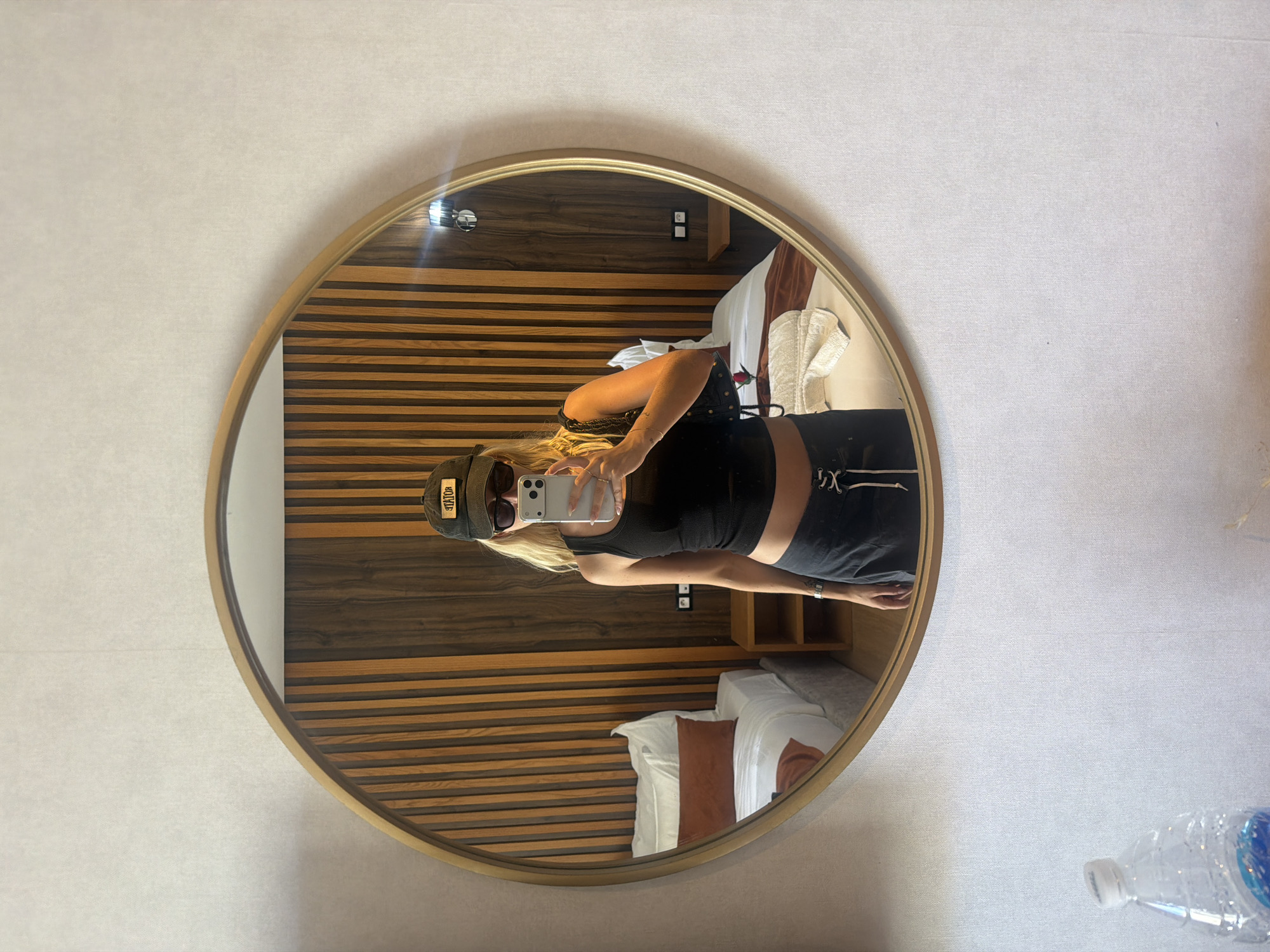 A blonde-haired woman wearing a black tank top, dark athletic shorts, sunglasses, and a baseball cap is taking a selfie in a circular mirror. The mirror hangs on a textured, light-colored wall. The reflection shows a headboard with alternating dark and light wood panels. A bed with white linens and brown accent pillows is partially visible. The location appears to be a room in a hotel or residence. The scene does not inherently provide information about the city of Kenitra, Morocco.