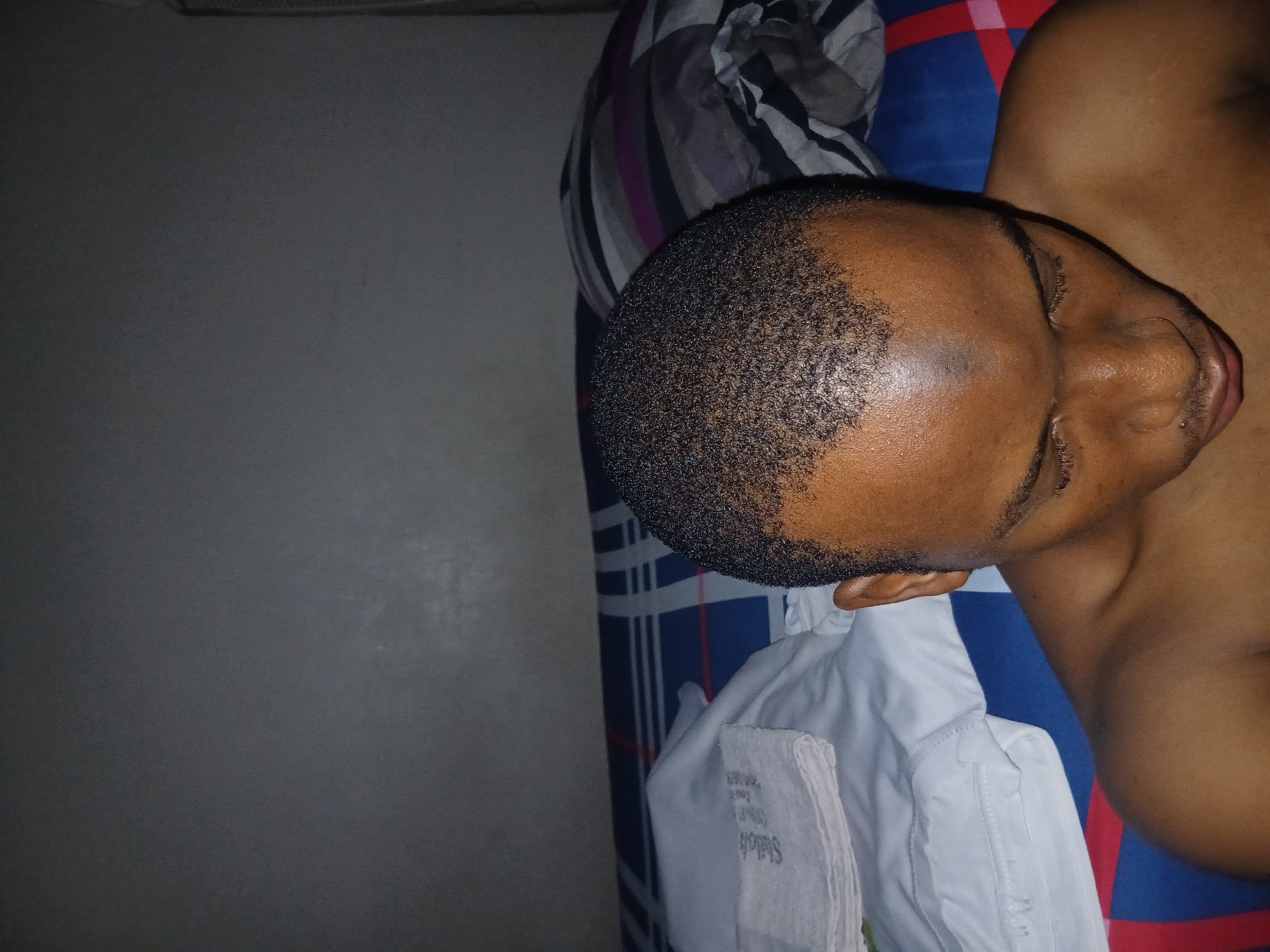 The image shows a close-up, overhead view of a dark-skinned individual with short, dark hair, looking downwards. The individual is shirtless, and their shoulders and neck are visible. The background consists of a bed with a blue, white, and red plaid patterned sheet, and a pillow with a black, white, and grey striped pattern. A white folded towel or cloth with faint writing is on the left side of the bed. The lighting is dim, suggesting an indoor setting, possibly a bedroom. The scene does not contain any elements that would allow for the specific identification of the city of Lagos, Nigeria.