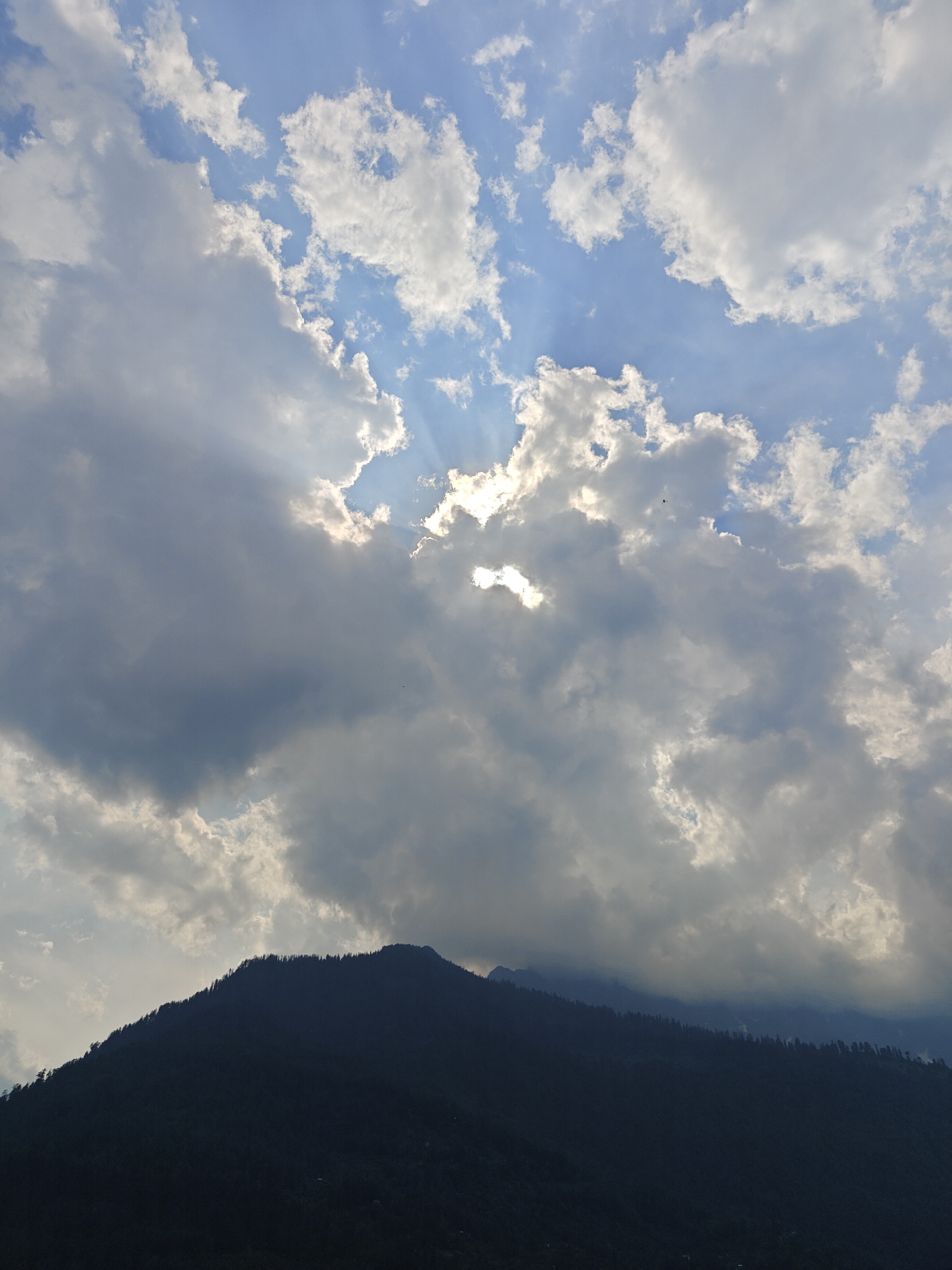 The image shows a silhouetted mountain range against a sky filled with dramatic clouds and sunbeams breaking through. The mountain is covered in dense, dark trees that create a textured silhouette against the lighter sky. In the sky, the sun is partially obscured by large, billowing clouds, creating rays of light that pierce through the overcast conditions. Some smaller, brighter clouds are scattered across the blue sky. The overall scene suggests a natural landscape, possibly taken during late afternoon or early morning when the sun's light is angled. The location is identified as Vashisht, India.