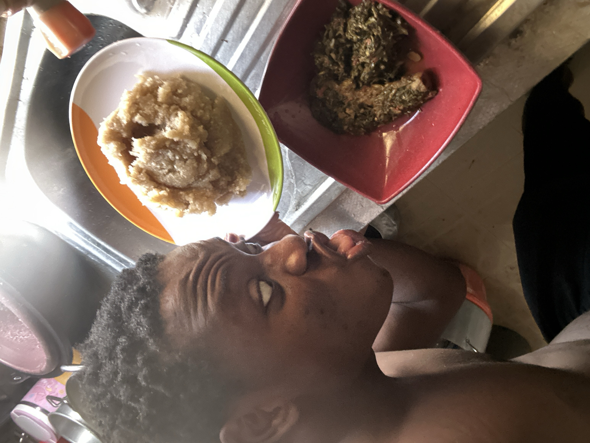 A dark-skinned person lies sideways in an unknown city, with two plates of food situated above them. One plate contains a portion of what appears to be stewed greens, and the other holds a lump of beige food. The person's face is visible, with an eye partially open and mouth slightly ajar. A stainless steel sink is visible in the background, along with the edge of a countertop.