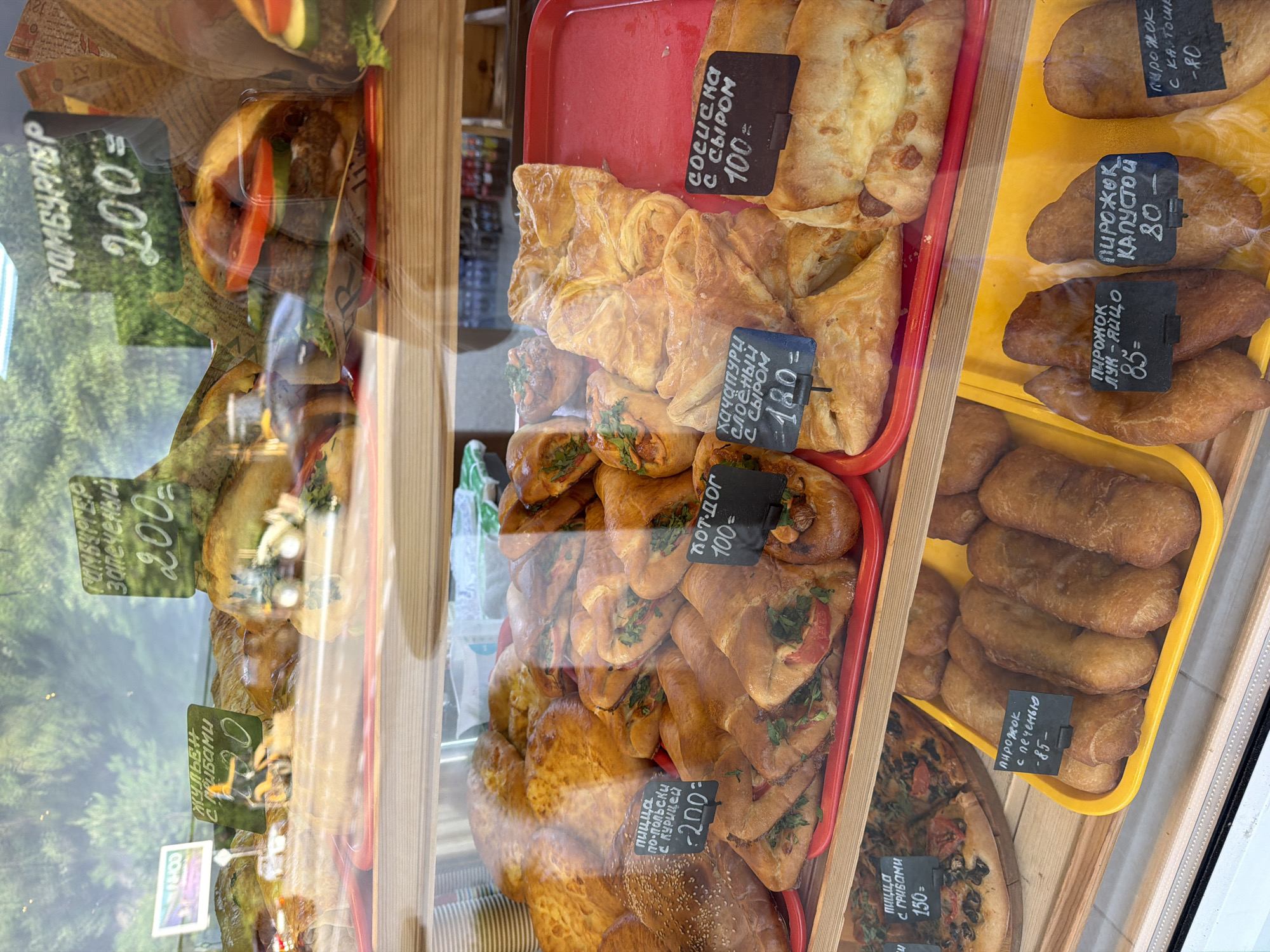 The image shows a bakery display case filled with various baked goods for sale. Items include pastries, pies with fillings such as meat, cabbage, and potato, pizza, and hot dogs. Each item has a small sign indicating its name and price in rubles, suggesting the location is in Russia. The writing on the signs is in Russian. Based on the products and pricing displayed, the picture appears to be taken in a city with a local bakery or shop. There is nothing in the image indicating that this is the city of Sochi. The food items are arranged on colored trays within the display case, which has a reflection of the outside environment, showing trees and possibly a building across the street.