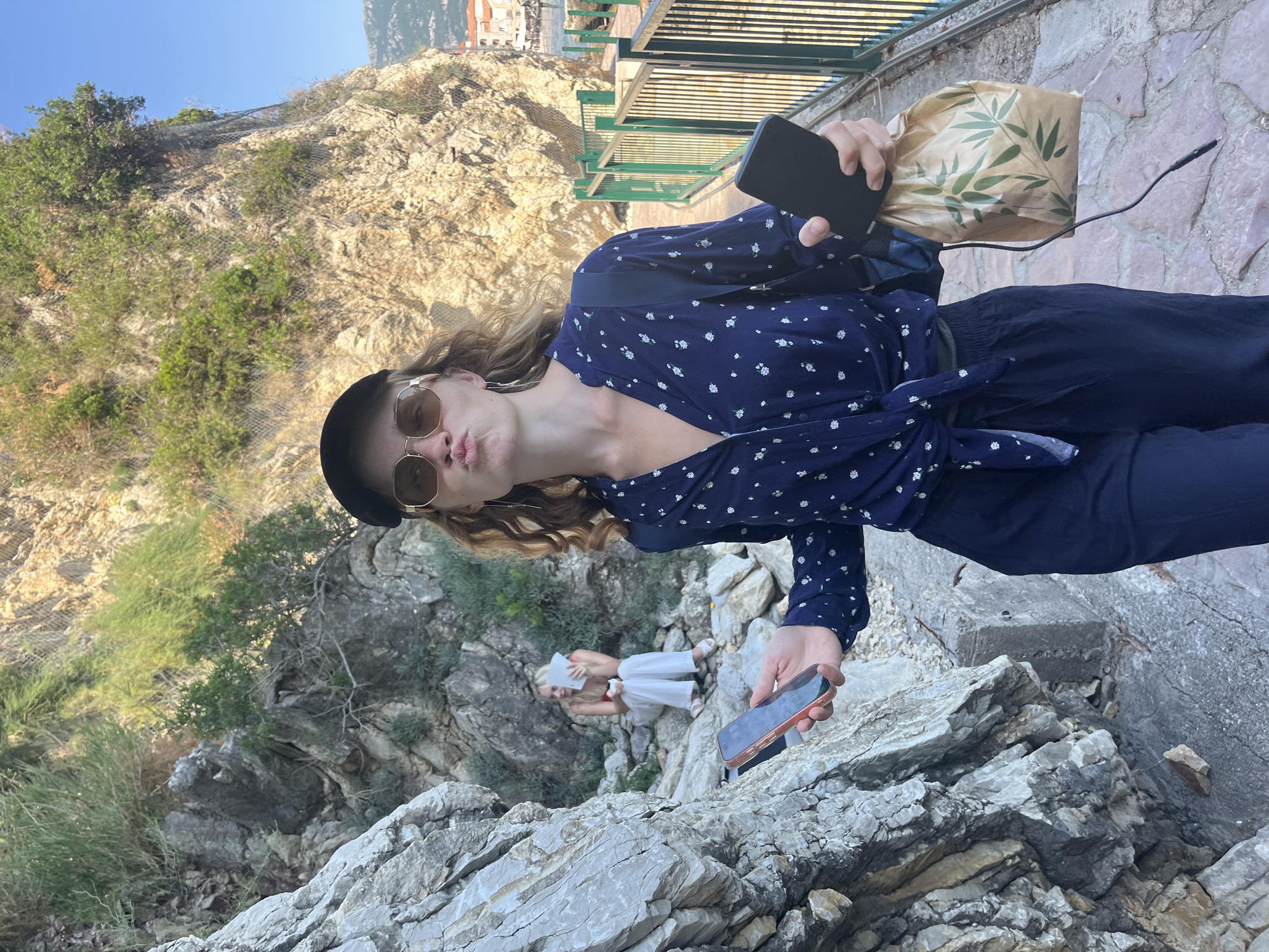 The image shows a young woman with blond curly hair, wearing a navy blue blouse with white floral patterns tied at the waist, navy pants, a black beret, and gold-rimmed sunglasses. She is holding a black smartphone in her right hand and a brown paper bag with green leaf designs in her left hand. She is making a kissing face towards the camera.
The woman is standing on a rocky area, with a steep hillside covered in vegetation behind her. The background also includes metal fencing and buildings suggesting an urban area.
A second person is visible in the background, seated on rocks and wearing white pants and a white top. They appear to be reading a book or using a tablet. Another phone is present on the rocks where the first woman is standing.
Based on the mountainous coastal landscape, the location is likely somewhere near the city of Budva, Montenegro.