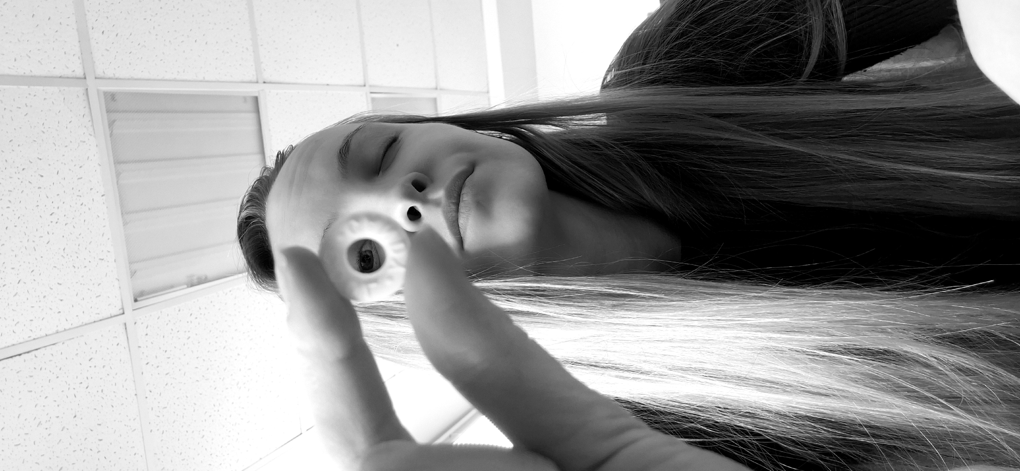 A black and white, close-up, upward angle image depicts a young woman holding a candy in front of her face. The woman has long hair that covers most of the right side of the image and extends towards the bottom. The ceiling in the background is a white, tiled drop ceiling. The person's eyes are closed and her mouth is slightly open. She is holding a white, ring-shaped candy between her index and middle fingers, positioned between her nose and the viewer, with her eye reflected in the candy's hole. The setting is most likely indoors, potentially a room with standard office-type suspended ceiling. There is nothing to indicate that this is located in Chaikovsky, Russia.
