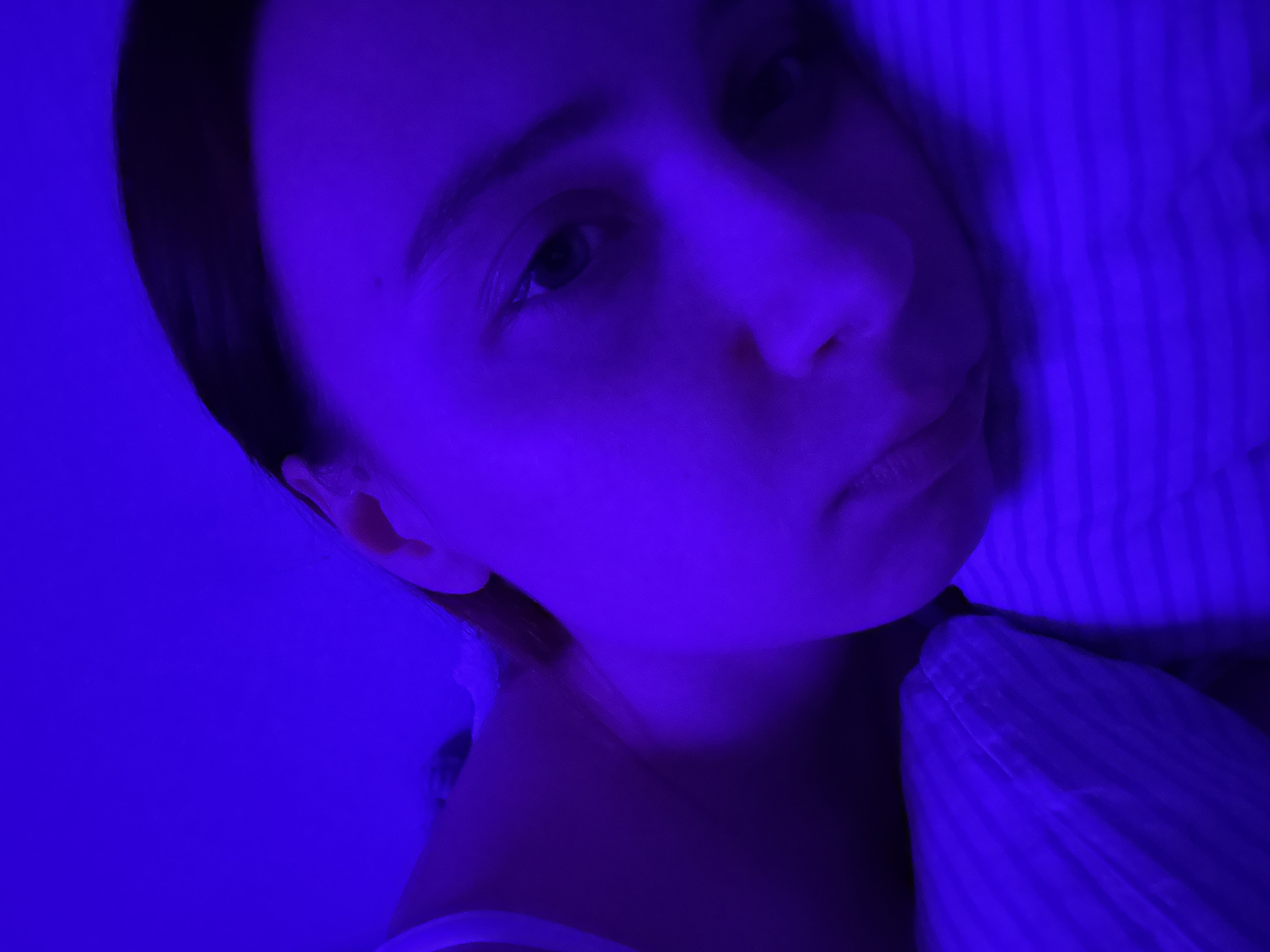 A close-up selfie of a young woman is illuminated by a strong blue light. The woman is lying down on what appears to be a striped pillow. Her face is centered in the frame, and her expression is neutral. Her hair is dark and pulled back from her face. Her right ear is visible. The background is heavily cast in the same blue light. The location in the city of Прикамский, Russia is not evident from the image.