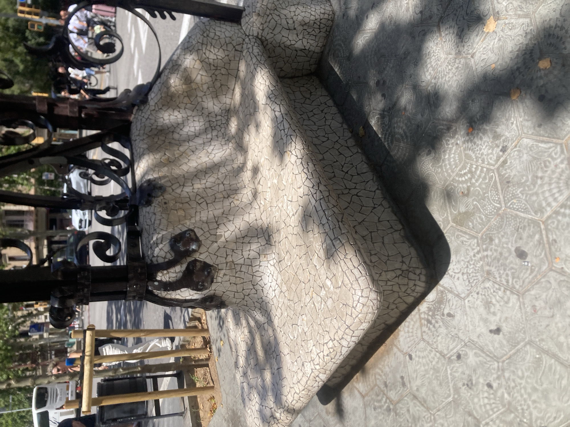 This is a medium-shot, eye-level, outdoor photograph of a bench in Barcelona, Spain. The bench is made of a mosaic of small, irregular white tiles and is curved, with a wider seat and a narrower backrest. It is supported by black metal decorative legs and a base covered in similar mosaic tiles. The bench is situated on a sidewalk made of hexagonal tiles, some of which have intricate patterns. Sunlight casts shadows across the bench and the sidewalk. In the background, a street scene is visible with people walking, vehicles, and buildings. A woman with blond hair is walking to the left of the frame, wearing white pants and a dark top, and a white bus is parked in the background. There are also some trees visible. The overall impression is a sunny day in a public urban space.