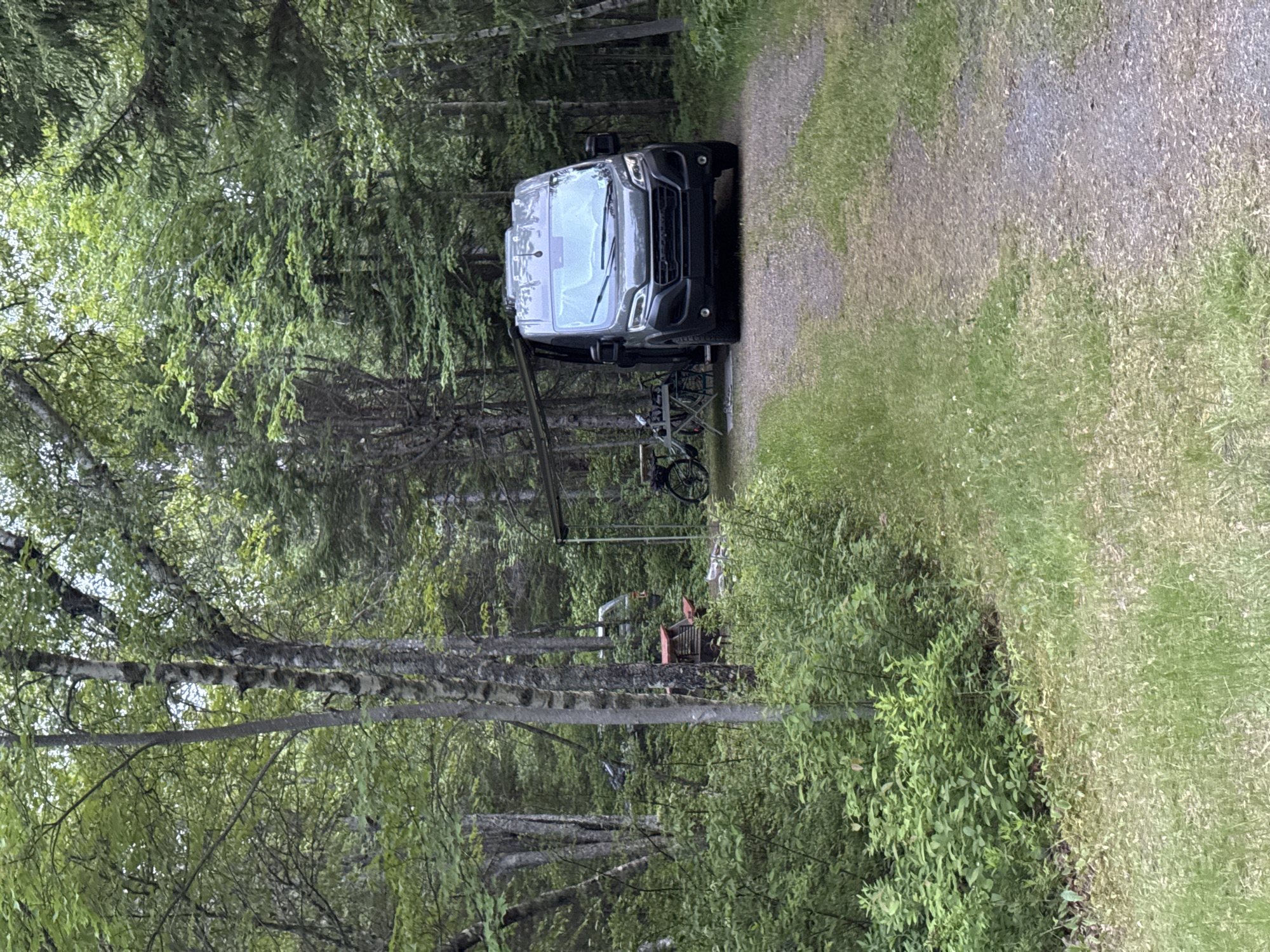A dark-colored camper van is parked on a gravel road in a wooded area. The van is facing forward, with its windshield and front grille visible. It appears to be a modern model with tinted windows. To the left of the van, a bicycle is mounted on a rack. A metal frame structure, possibly a canopy or awning, is set up behind the van. In the background, dense green foliage and tall trees create a natural, secluded setting. The ground is covered in gravel and grass, suggesting a campsite or a rural road. The scene is likely outdoors, with natural lighting, suggesting daytime. There is no visible text in the image. The overall impression is one of a peaceful, natural environment.