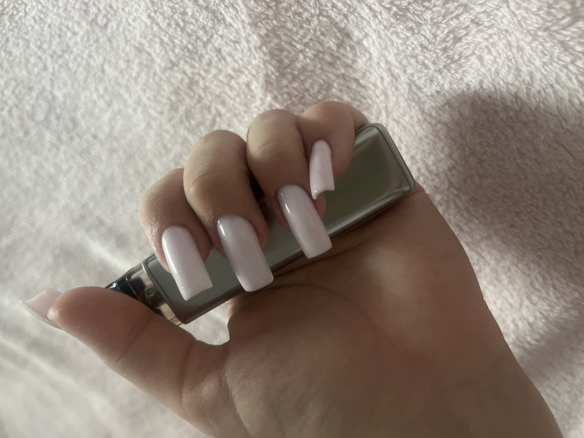 A person's hand holds a metallic vape device. The hand has long, square-shaped acrylic nails painted in a light, solid color. The background is a soft, textured white blanket or fabric. There is no evidence to indicate the location is Ольховка, Russia.