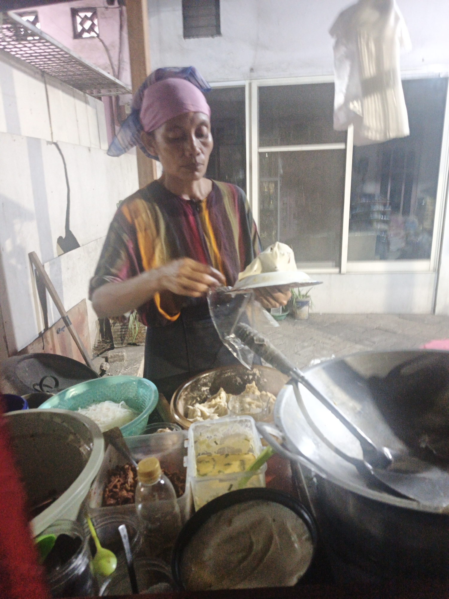 A woman wearing a pink headscarf and a striped shirt is preparing food outdoors in Surabaya, Indonesia. She is standing at a makeshift food stall with various ingredients and cooking utensils. To her left is a green bowl filled with white noodles. In front of her, she holds a white plate with food, and to her right is a large metal wok. The background shows a building with windows and a shelf with decorative objects. The overall impression is of a street food vendor at work.