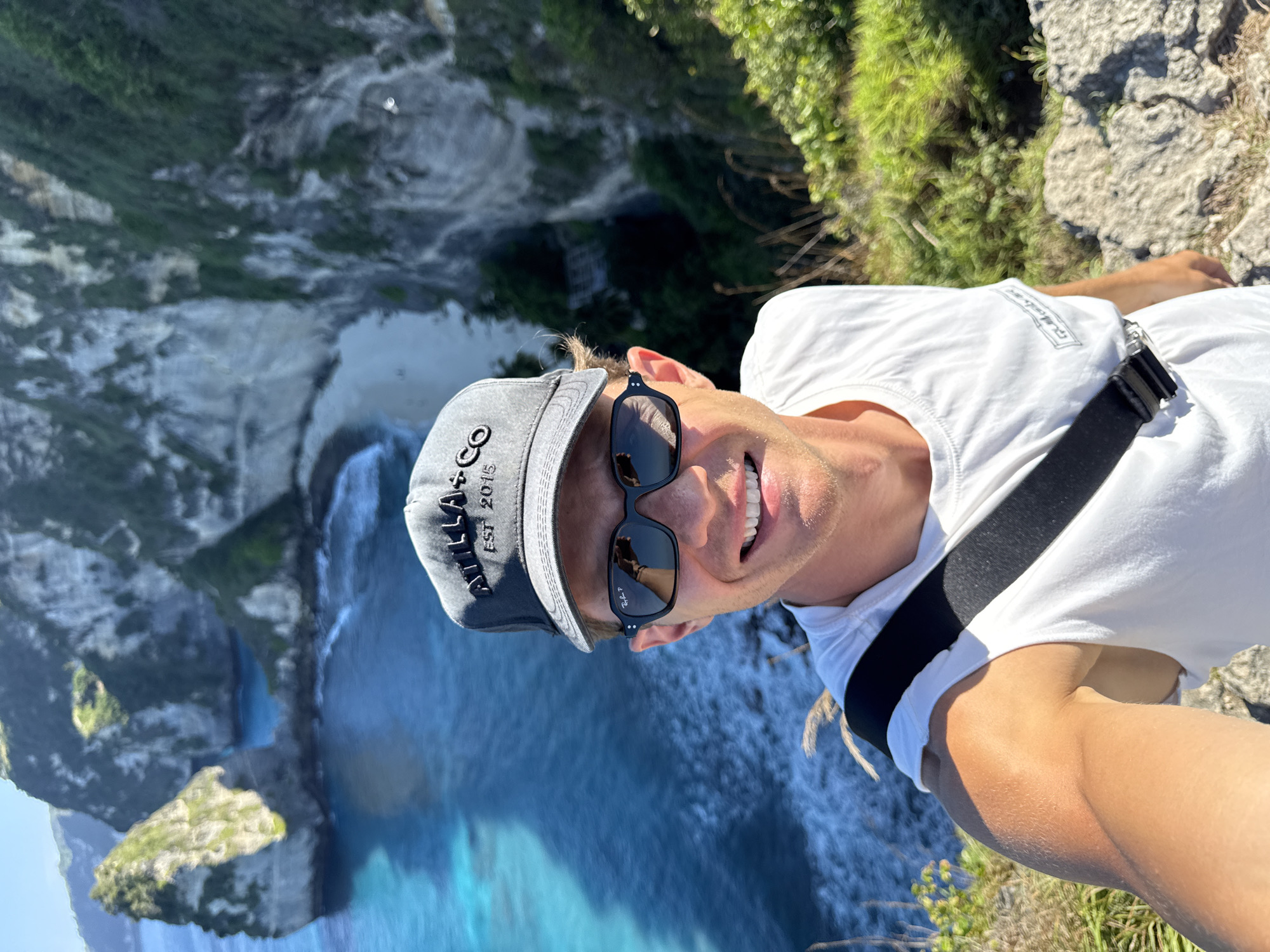 In the city of Nusa Penida, Indonesia, a man wearing a grey baseball cap and sunglasses takes a selfie from a cliff overlooking a turquoise ocean and a beach. The man is wearing a white t-shirt and has a black strap across his chest. The background features dramatic cliffs with lush green vegetation and the clear blue waters of the ocean, with a small beach visible below. The overall scene depicts a scenic coastal landscape.