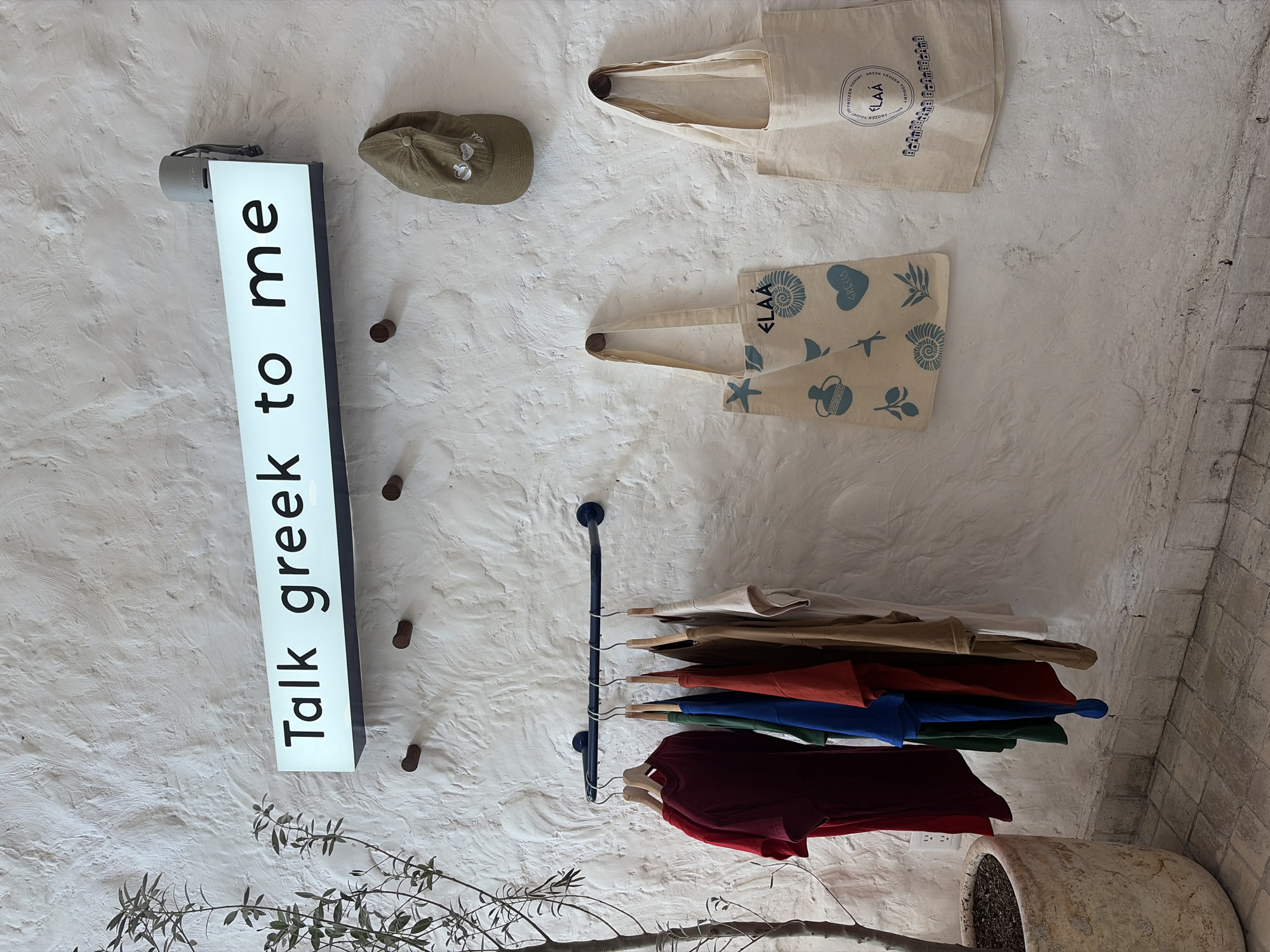 This image shows a wall display in a store in Unknown, Unknown. The wall is textured and painted white. On the wall are a sign that says "Talk Greek to me," a tan baseball cap, two tote bags with Greek-themed designs, and a rack with several colorful shirts hanging on it. A potted plant is at the bottom right of the frame.