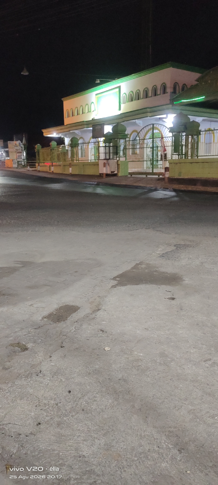 The image depicts the exterior of a building, likely a mosque, at night. The building features a white facade with green trim and arched windows. A fence surrounds the building, and there is a flag with red and white stripes visible. The foreground shows a wet asphalt road with some puddles. The bottom left corner of the image contains a watermark indicating it was taken with a "vivo V20" phone by "ella" on "25 Agu 2026 20:17". The scene is likely located in Wates, Indonesia, given the presence of the Indonesian flag. The lighting suggests the image was taken at night, with artificial lights illuminating the building. No people are visible in the frame.