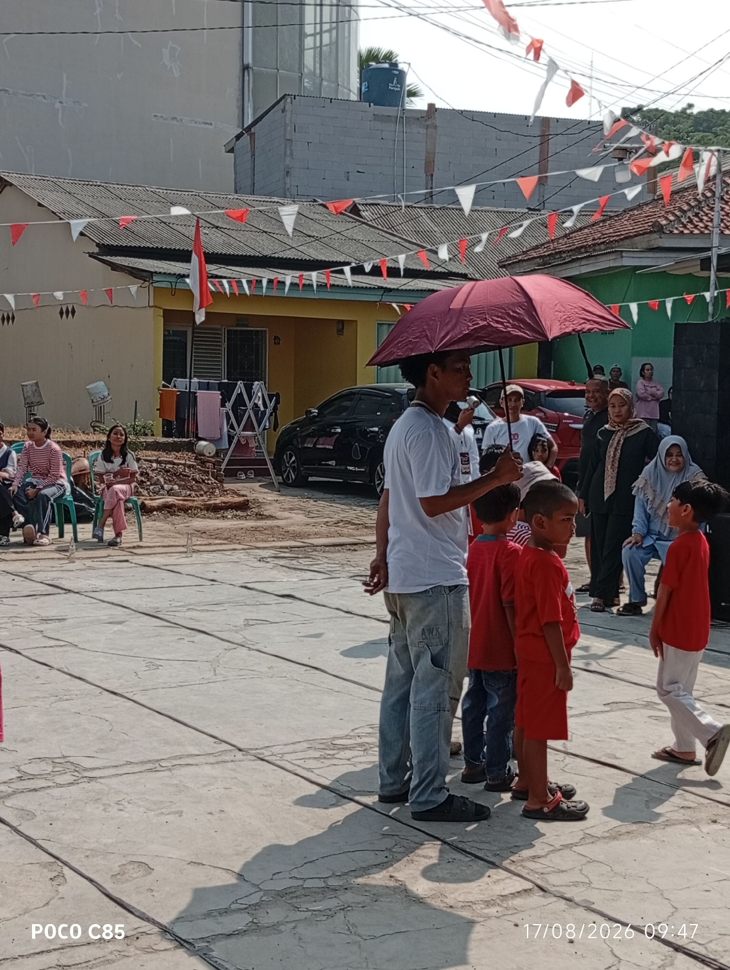 In a street setting in the city of South Tangerang, Indonesia, a group of people, predominantly children, are gathered outdoors. The scene is decorated with red and white triangular flags strung across wires, suggesting a festive or commemorative occasion. A man holding a maroon umbrella stands in the foreground, facing a group of children who are looking in his direction. Some of the children are wearing red shirts. In the background, there are houses with tiled roofs and a black car parked on the street. Several adults are also visible, some observing the gathering. The ground is paved with cracked concrete. The timestamp "17/08/2026 09:47" is visible in the lower right corner of the image.