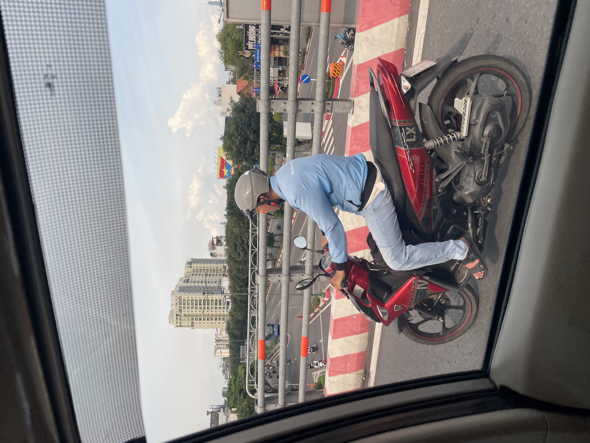 The image depicts a person riding a red Yamaha motorcycle on a bridge in Thủ Đức, Vietnam. The rider is wearing a light blue long-sleeved shirt, blue jeans, a gray helmet, and black sandals. The motorcycle has black wheels with a red rim accent. In the background, there is a multi-lane road with other vehicles, including a blue bus and several other motorcycles. Buildings and trees are visible along the sides of the road. The bridge is lined with a barrier of alternating red and white concrete blocks and metal railings with orange accents. The sky is partly cloudy. The perspective suggests the photo was taken from inside another vehicle.