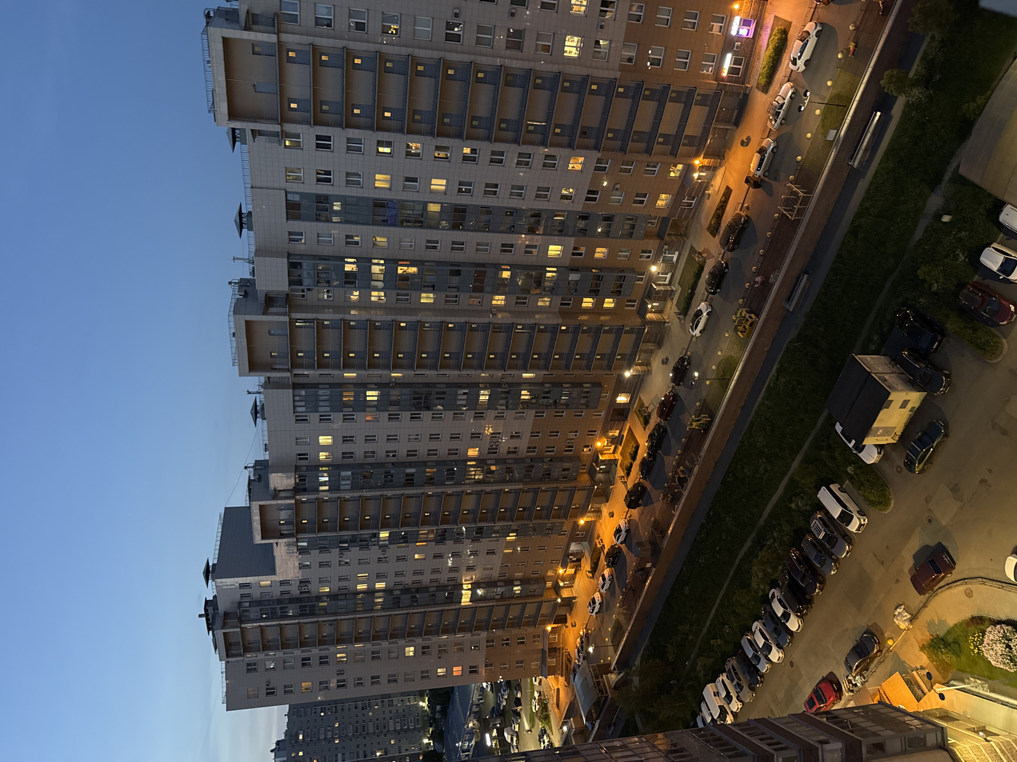 This is a high-angle, full shot of a modern, multi-story residential building complex at dusk in an urban environment. The sky is a deep blue with no visible clouds. The building is tall and appears to be a single, continuous structure with numerous balconies and windows, many of which are illuminated, indicating occupancy. The facade of the building is a combination of light-colored materials, possibly concrete or stone, with darker accents and large glass panels. There are several other residential buildings visible in the background, appearing to be of similar construction. In the foreground and along the side of the main building, there is a paved area with a parked row of cars, including sedans and SUVs, in various colors such as white, black, silver, and red. A strip of green grass separates the parking area from a sidewalk or pedestrian path. Further in the background, more cars are parked in what appears to be a parking lot. The overall scene depicts a typical evening in a residential area of a Russian city, likely Moscow or Saint Petersburg, given the architectural style and the presence of a large apartment complex.