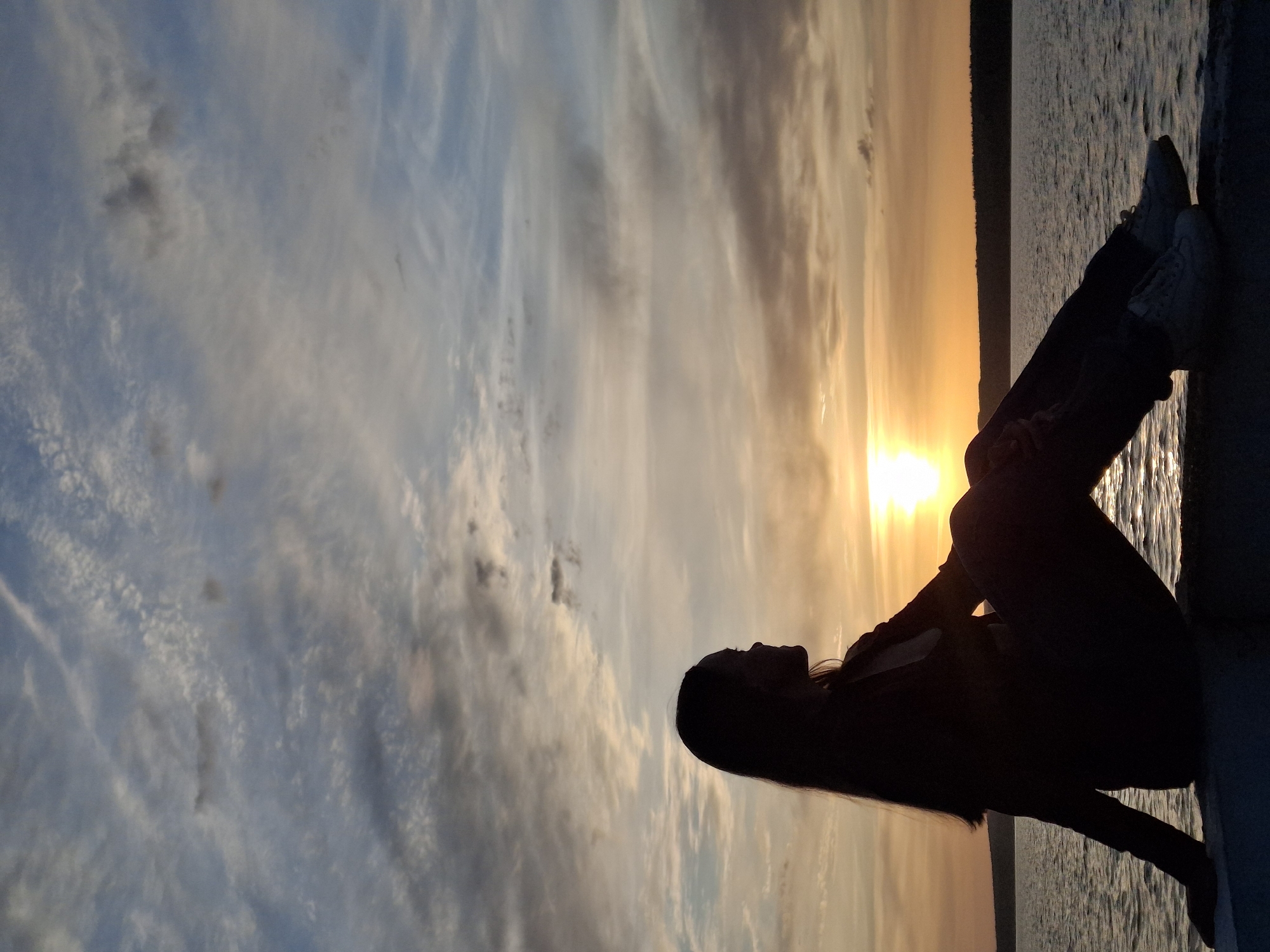 A silhouette of a person is positioned horizontally along the right side of the image, set against a sunset-tinged sky. The person is sitting with legs crossed at the ankle and feet extended, resting on a concrete ledge. The sky is filled with a mix of blue and orange hues, suggesting either sunrise or sunset. The bright sunlight is visible at the horizon. A dark strip of land or trees is seen on the horizon as well. The location is in Chaikovsky, Russia.