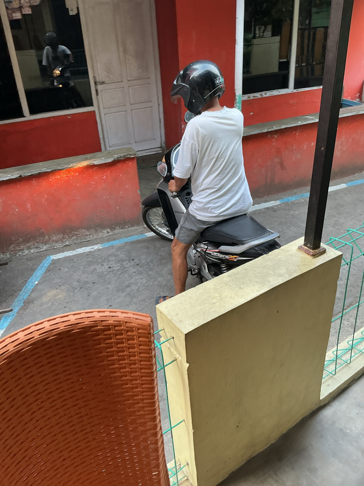 In the city of Magelang, Indonesia, a person wearing a black helmet and a white t-shirt is seated on a motorcycle. The individual is facing away from the camera, with their back to the viewer. They are wearing gray shorts and flip-flops. The motorcycle appears to be parked or stopped. In the background, a red building with a white door is visible, and a reflection in a window shows another person on a motorcycle. The ground is paved with concrete and marked with blue and white lines, possibly indicating parking spaces. In the foreground, a portion of a brown, woven chair and a light yellow concrete structure are present. A green metal fence is also visible.