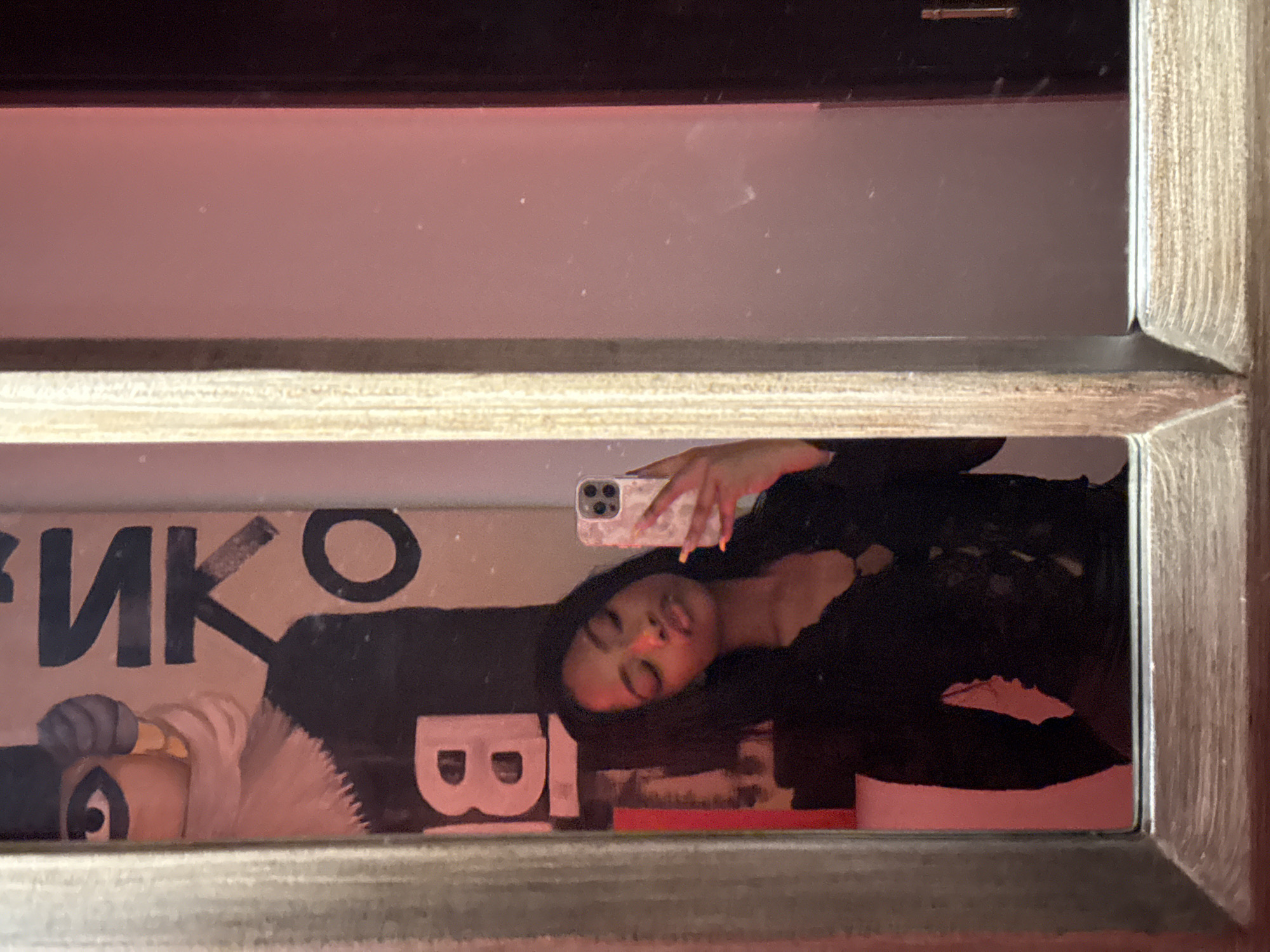 A woman is taking a selfie in a mirror. The reflection also shows a poster with the Cyrillic letters "ИКО" and the letter "B". The location is unknown; there is no information to ascertain the city of Moscow, Russia.