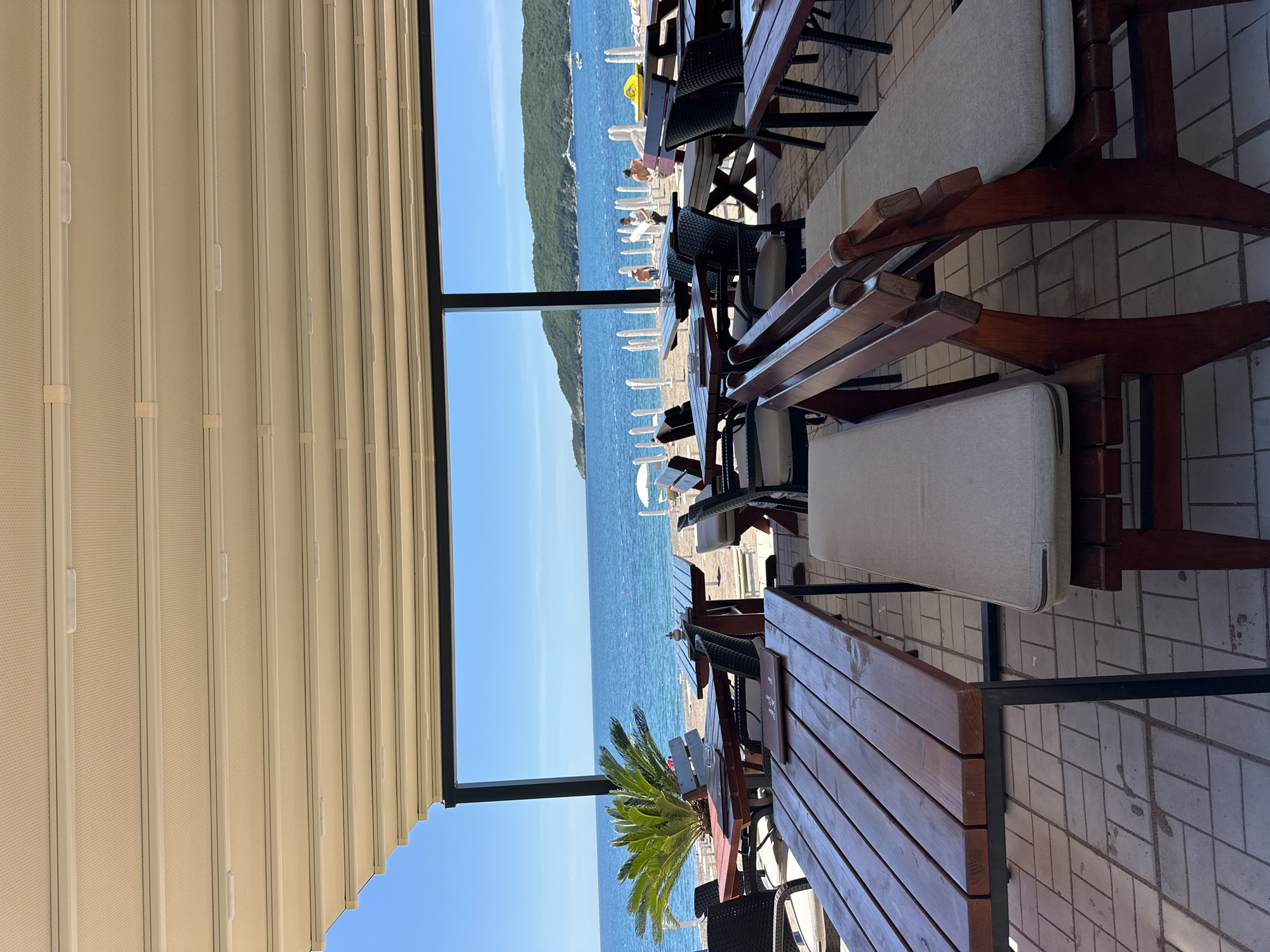 The image shows an outdoor seating area of a cafe or restaurant in Boreti, Montenegro, overlooking the Adriatic Sea. Several wooden tables and benches with light-colored cushions are arranged on a tiled patio. The tables appear to be set for customers. Through a large, rectangular opening framed by a black structure, the view extends to a beach with white umbrellas and people enjoying the sunshine, as well as the blue water of the sea, with a lush, green, mountainous coastline in the distance. A retractable awning provides shade over the seating area. A small palm tree is visible near one of the tables. The sky is clear and blue.