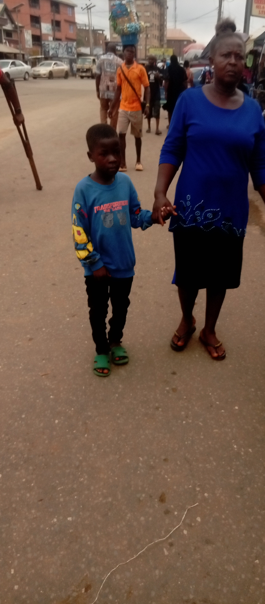 A full outdoor shot shows a young boy and an older woman holding hands while walking on an asphalt road in the city of Unknown, Unknown. The boy, facing forward, wears a blue sweatshirt with "Transformers: The Game" on it, black pants, and green sandals. The woman, to his right, is in a blue top and black skirt with flip-flops.
In the background, several people are walking; one man carries a large, colorful load on his head. A few parked cars are visible along the roadside, and buildings are in the distance. A long pole stands on the left edge of the frame. The scene appears to be on a day with overcast skies. A thin string is on the asphalt surface in the foreground.