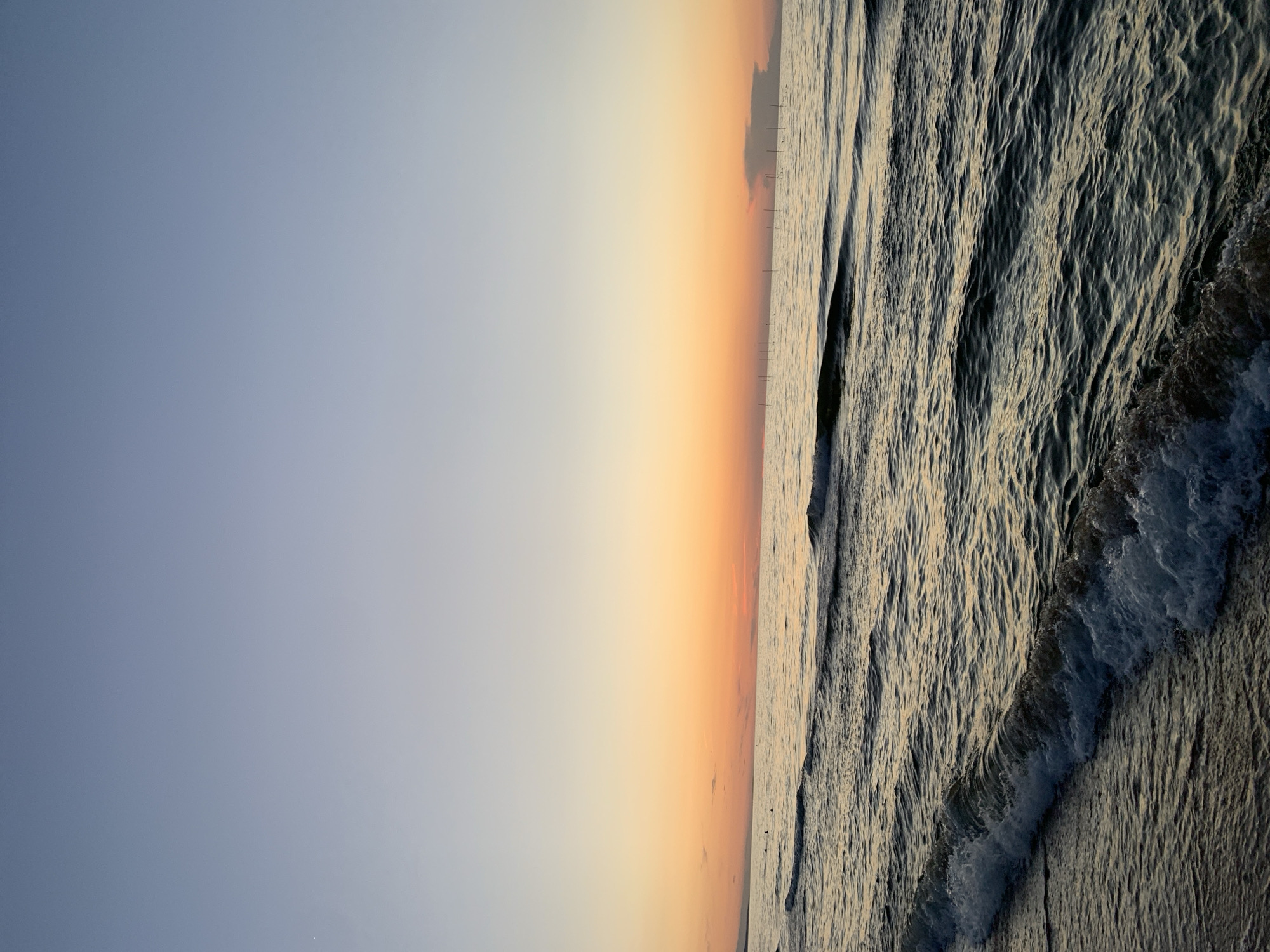 The image captures a serene sunset over the sea at Golubitskaya, Russia. The sky transitions from a clear, pale blue at the top to a vibrant orange and yellow gradient near the horizon, with a few faint clouds. The sea is calm, with gentle waves lapping at the shore. In the distance, a line of thin vertical structures, possibly fishing poles or markers, stretches across the water. A small, dark cloud formation is visible on the right side of the horizon. The foreground shows the sandy shore with the edge of an incoming wave, its white foam dissipating on the wet sand. The overall atmosphere is peaceful and tranquil.
