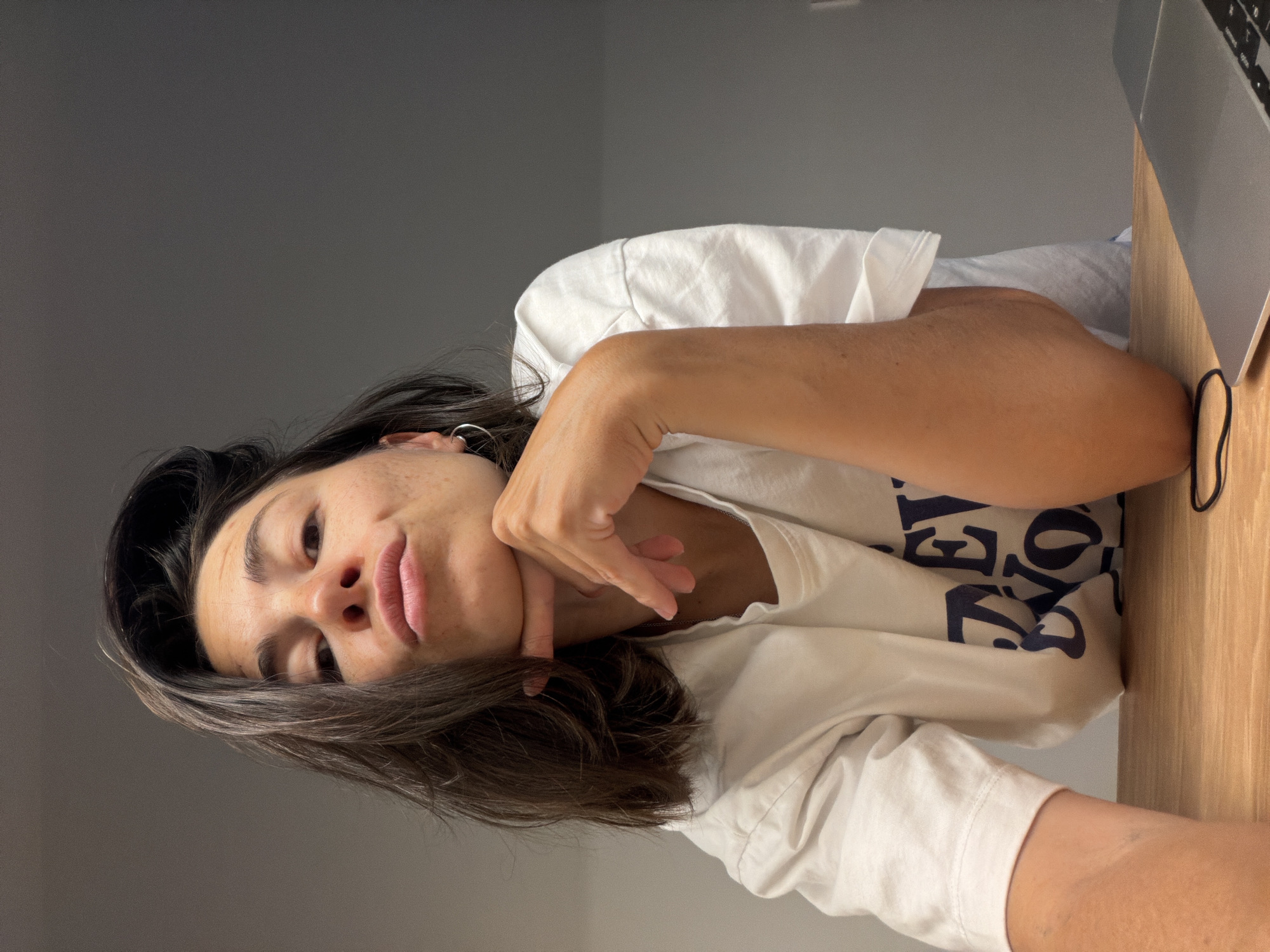 A woman is indoors, propping her chin with her hand and gazing toward the camera. She has dark hair and fair skin. She is wearing a white t-shirt with blue lettering. To her right is a laptop computer on a light-colored wooden surface. The wall behind her is a solid grey color. No location is given, so I am unable to describe the scene in the city of Budva, Montenegro.