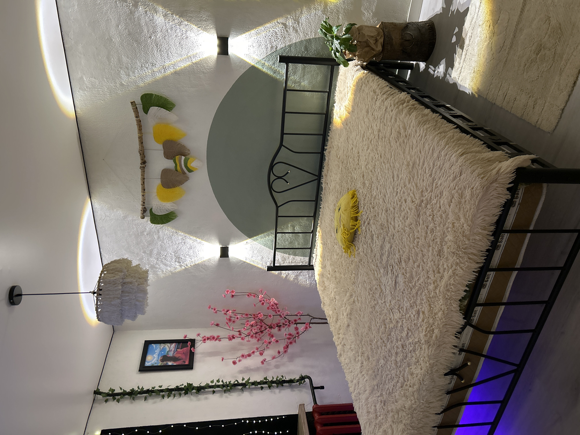 The image depicts a bedroom. In the center of the room is a bed with a plush, cream-colored comforter. A yellow decorative pillow rests on the bed. The bed frame is black metal with a heart-shaped detail on the headboard.
On the wall behind the bed, there are two wall-mounted lights that cast beams of light upwards and downwards. Above the lights, a decorative wall hanging made of driftwood and colorful felt leaves is displayed. The wall has a textured finish and is painted white with a light green arched section behind the bed.
To the left of the bed, there is a tall, artificial pink cherry blossom tree. To the left of the tree, a framed picture hangs on the wall, depicting a person looking out at a seascape. A strand of artificial ivy is draped along the upper left corner of the wall. On the far left, a red radiator and part of a window with string lights are visible.
To the right of the bed, a small potted plant sits on a rustic wooden stand. A cream-colored rug lies on the floor to the right of the bed, with a golden light illuminating a portion of it. Blue LED lights are visible beneath the bed frame.
The overall impression is that of a decorated, cozy bedroom. There are no people visible in the image. The scene is indoors, and there is no indication of the specific location within the city of Bălți, Moldova, beyond it being a bedroom.