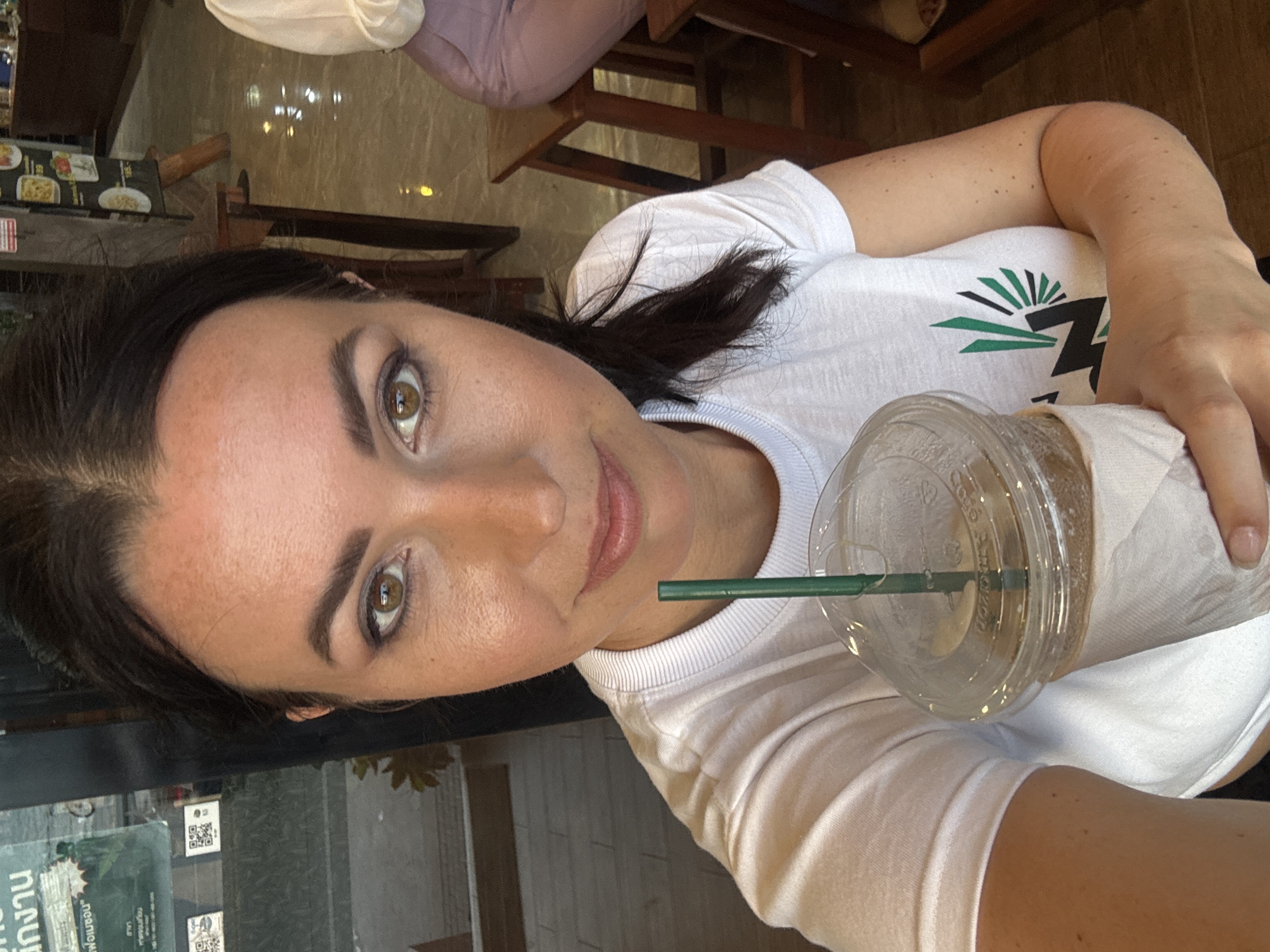 A woman is captured in a close-up, slightly tilted selfie, holding an iced beverage in a clear plastic cup with a green straw. She has dark hair, light brown or hazel eyes, and a subtle smile. Her left arm is visible, showing some freckles. She is wearing a white, short-sleeved t-shirt featuring a distinctive logo on the chest: green sun-like rays above a green horizontal shape and a bold black, stylized "Z" or abstract letter.
The setting appears to be an indoor cafe or restaurant, indicated by wooden furniture, tables, and reflections in what seems to be a window or polished wall behind her. The lighting suggests it is daytime. In the background, a menu or display with images of food items is visible.
Notable details include reflections in the background that contain Thai script, such as "บริการ" (service) and "งาน" (work/job), along with QR codes and part of a sign that reads "พัทยา" (Pattaya), confirming the location as Pattaya City, Thailand. The woman's expression conveys a sense of pleasantness or contentment. The iced drink is wrapped in a white paper sleeve.