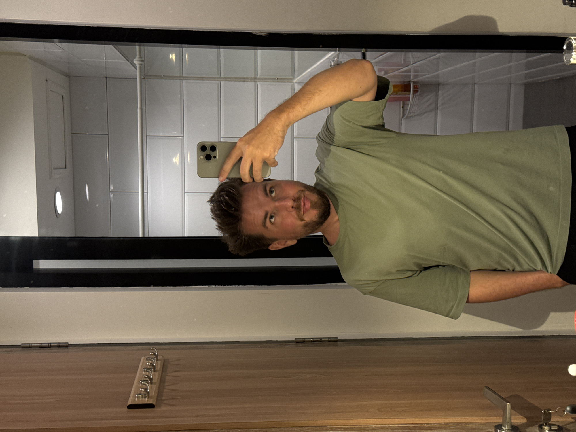 A Caucasian male with short brown hair and a beard is taking a selfie in a bathroom mirror. He is wearing a green t-shirt and holding an iPhone. The bathroom has white tiled walls and a wooden vanity with a row of hooks. The location is likely in North Kuta, Indonesia, based on visual cues.