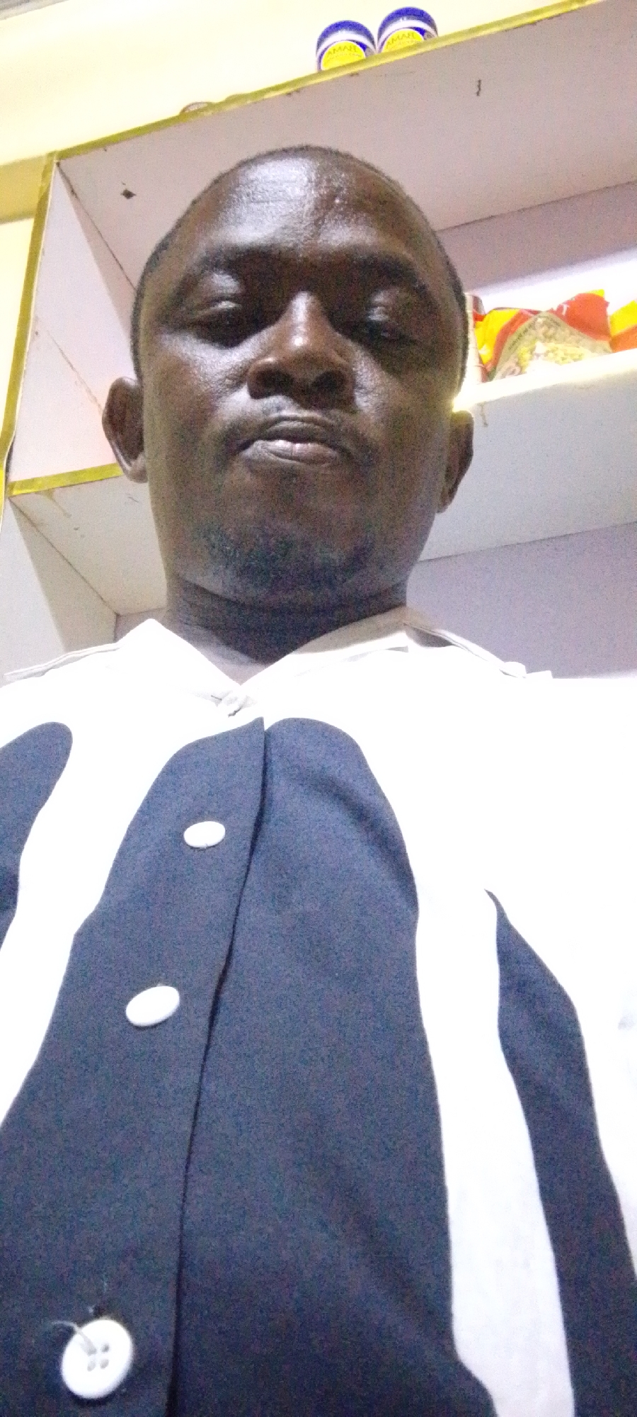 A dark-skinned man with a shaved head is facing the camera. He has a serious expression. He is wearing a black and white patterned shirt with buttons. Behind him is a white shelf. On the shelf are various containers. The location is likely indoors, possibly a residence in Adankolo, Nigeria, although specific location indicators are absent.