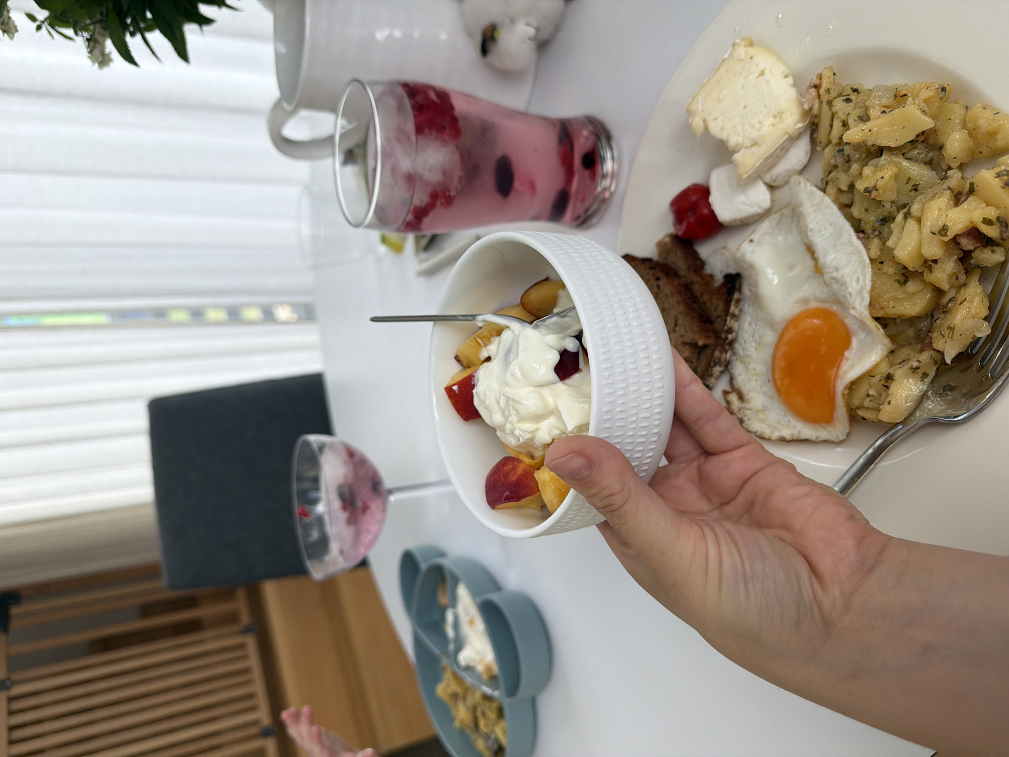 A high-angle, medium shot captures a white table laden with breakfast items. In the foreground, a person's hand holds a white bowl filled with yogurt and fruit. To the right, a glass of pink beverage with berries and ice sits next to a white teapot. A plate in the lower right contains a fried egg, potatoes, bread, and cheese. In the background, a baby gate and a chair are visible. The scene is set indoors with natural light filtering through sheer curtains.