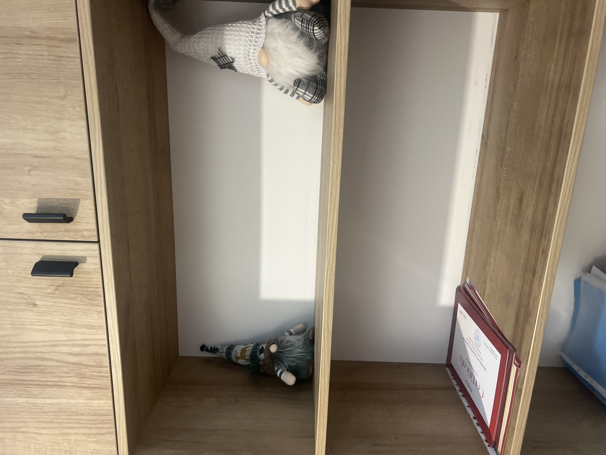 The image shows the interior of a wooden shelf unit. The top shelf of the unit is empty except for two black handles on the cabinet doors above. The second shelf contains two decorative gnomes. One gnome is on the left side of the shelf, wearing a green and white striped hat with a deer pattern and a green outfit with a brown beard. The other gnome is on the right side of the shelf, wearing a white textured hat with a plaid star, a white and gray striped shirt, and plaid pants, with a large white beard. The third shelf has a stack of certificates with red borders. The bottom of the unit is partially visible and contains some blue plastic folders. The overall environment appears to be indoors, possibly a home or office.