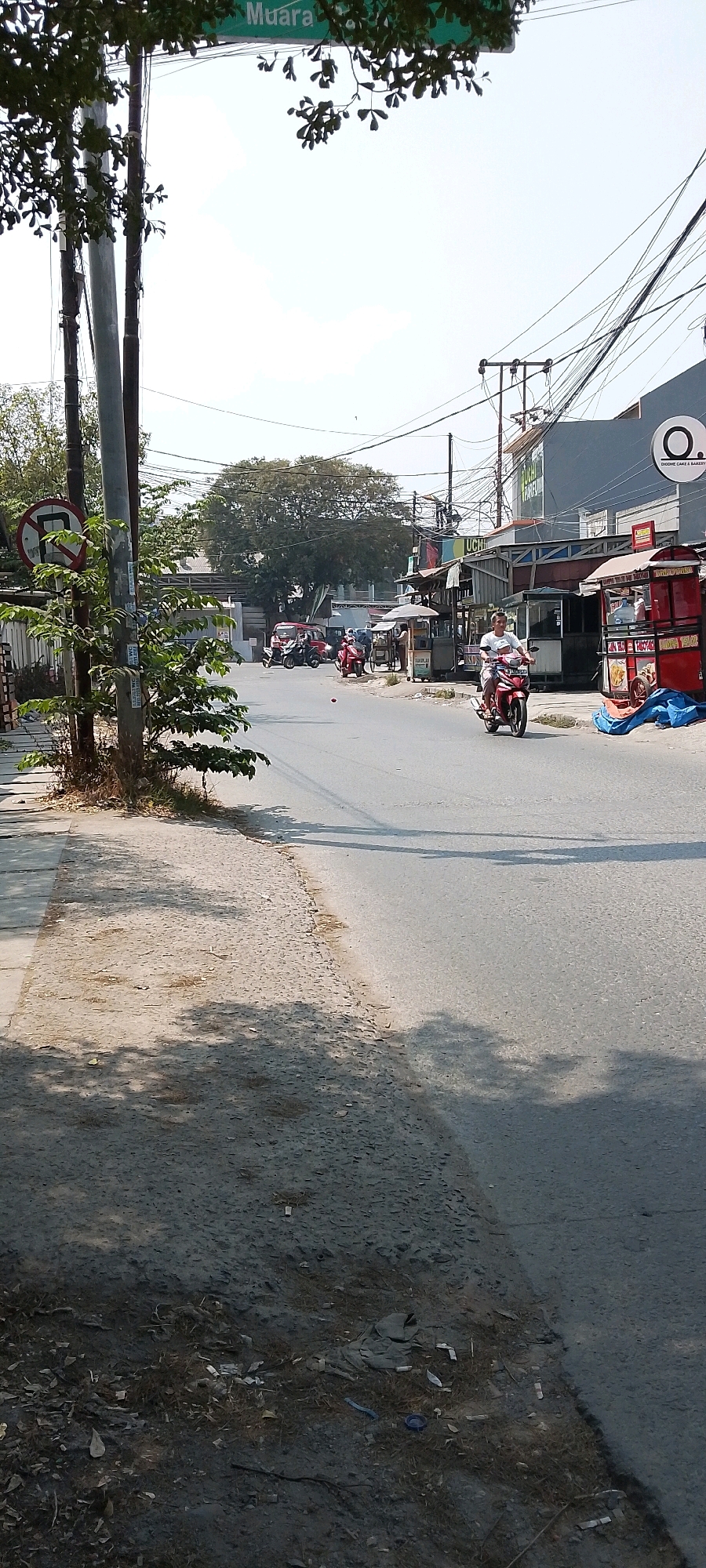 The image depicts a street scene in Babelan, Indonesia. A person is riding a red motorcycle on the right side of the road. Several food stalls are visible on the right side of the street. A large tree is in the background, and a street sign reading "Muara" is visible at the top of the image. There are also utility poles and wires overhead. The street appears to be paved, and there is a sidewalk on the left side of the image. The sky is clear and bright, indicating daytime.