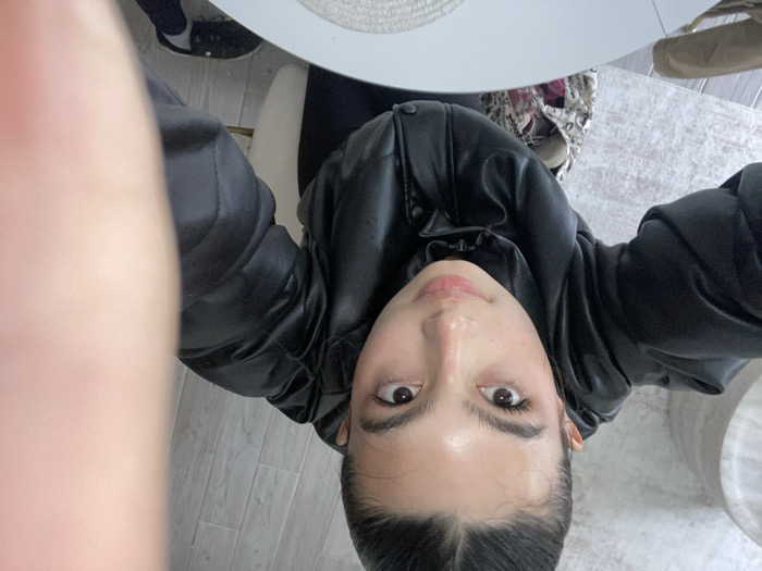 From a high angle, a young woman with fair skin, dark hair pulled back, and visible long eyelashes gazes upwards. She wears a black leather jacket, and her body is angled slightly to the left. A round table with a neutral-colored top is partially visible at the top of the frame. A light-colored chair and a rug are in the background. A person's shoe can be seen at the edge of the frame. A large, blurry object dominates the left side of the image.