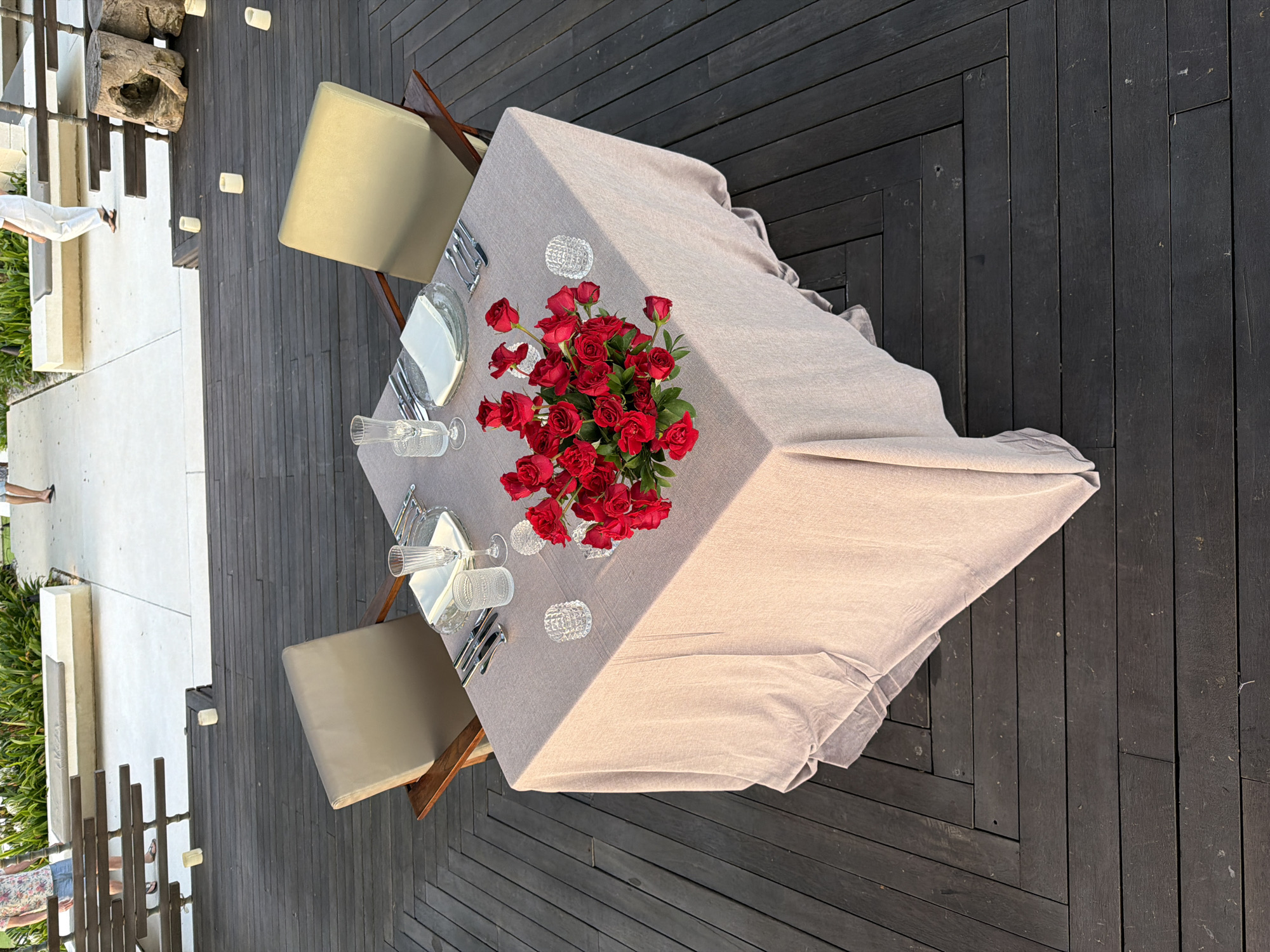 The image shows a romantic dinner setting on a wooden deck in Pecatu, Indonesia. A square table is set for two, covered with a light-colored tablecloth. A large bouquet of red roses serves as the centerpiece. Each place setting includes a plate, silverware, multiple glasses, and a decorative candle holder. Two chairs, upholstered in a light beige fabric, are positioned on opposite sides of the table. The deck is constructed of dark wood planks, and the surrounding area includes greenery and a white stone structure. People are visible in the periphery near the building and railing. Small white lights are placed in intervals along the walkway.