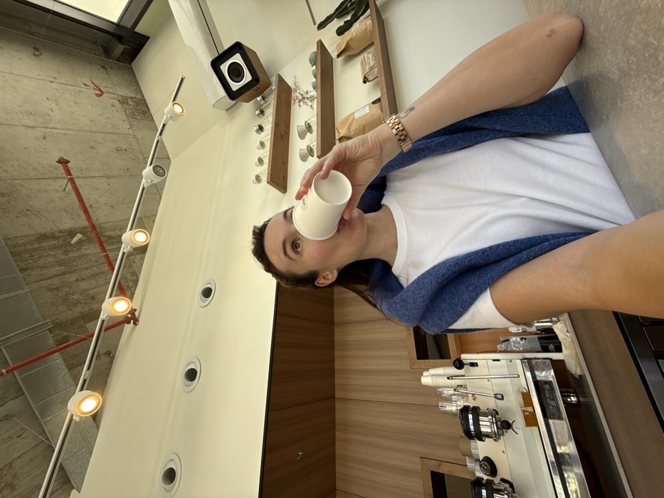 A female individual is depicted in an indoor setting, identified as a café, specifically Sunday Project Coffee in Tel-Aviv, Israel. The individual, seen from the chest up, has dark hair pulled back and is wearing a white t-shirt with a blue knitted sweater draped over her shoulders. She is actively drinking from a white, disposable paper cup, with her head tilted slightly upwards. A rose gold-toned wristwatch is visible on her left wrist. Her left arm is extended towards the camera, indicating a self-portrait.
The environment features a light gray countertop in the foreground. Behind the individual, a commercial espresso machine with multiple group heads and steam wands is present on the left. The background includes light wood paneling and a white wall section. Wooden shelves are mounted on the right wall, displaying several glass pour-over coffee vessels on the upper shelf and multiple brown paper bags, one bearing "SUNDAY," along with a small cactus plant on the lower shelf. The ceiling consists of exposed concrete with visible red piping/conduits and a track lighting system featuring several illuminated white spotlights. Additional recessed spotlights are visible in a dropped ceiling section. An air conditioning unit and a square, wooden-encased speaker or camera are mounted high on the right wall/ceiling. The lighting suggests an indoor daytime scene.