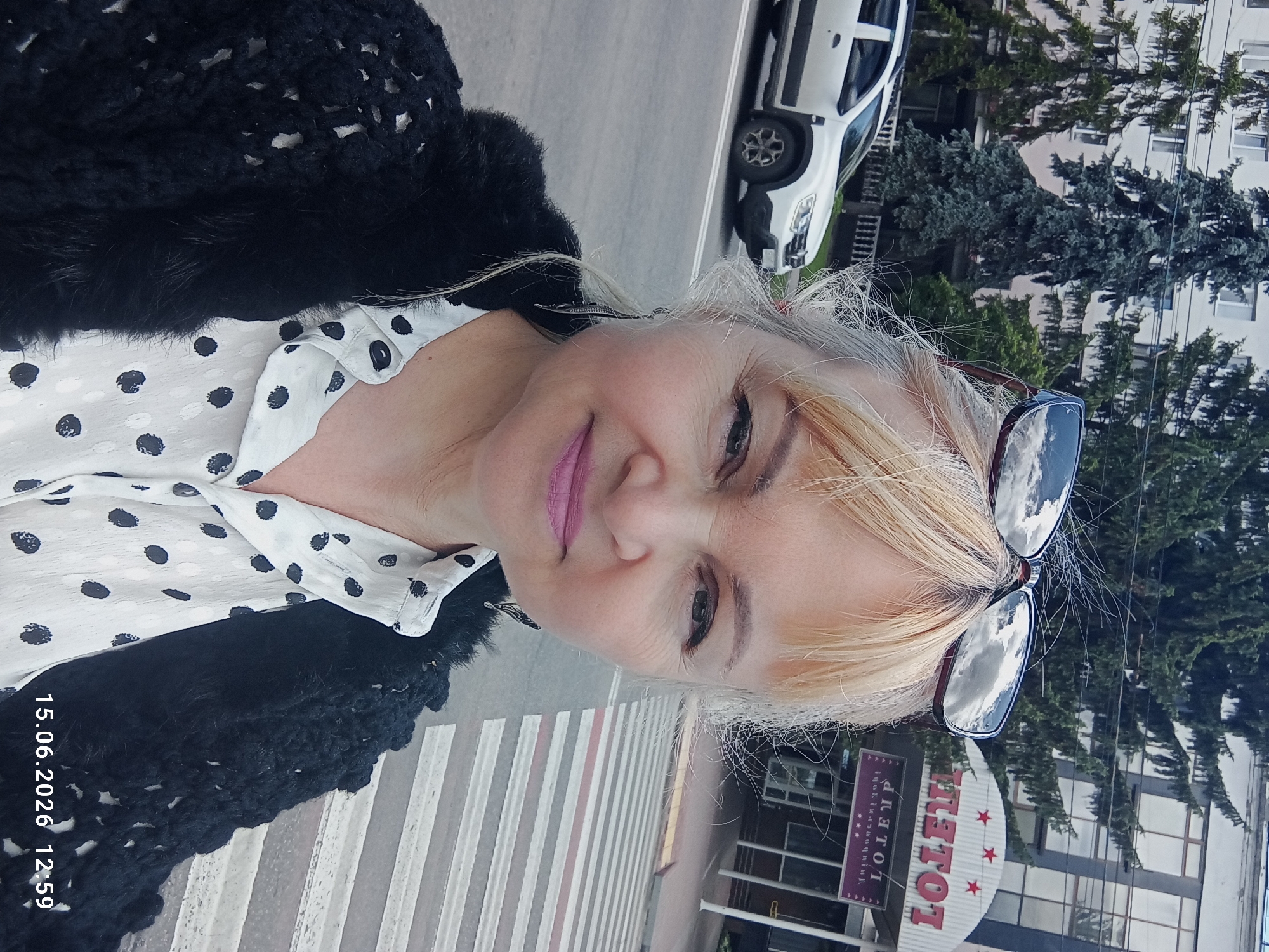 A woman with blonde hair is taking a selfie outdoors. She is wearing a black knitted sweater over a white polka-dot shirt. Her sunglasses are perched on her head, reflecting the sky. Behind her, a white car is parked on the street, and a building with a sign that reads "TOTEM" is visible. A pedestrian crossing is in the foreground. The date and time stamp on the image indicates 15.06.2026 at 12:59.