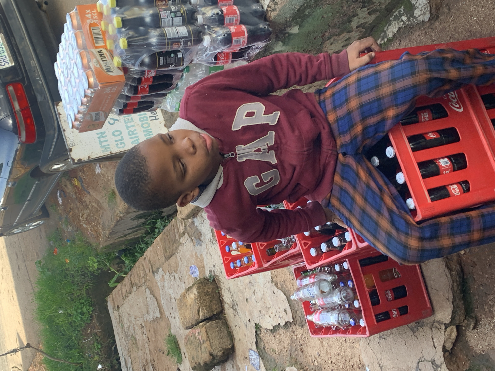 In a tilted outdoor scene in Jos, Nigeria, a young boy, appearing to be around 8-10 years old, is the central subject. He is wearing a maroon hooded sweatshirt with the "GAP" logo prominently displayed and blue and orange plaid trousers. The boy is seated amidst stacks of beverages, primarily Coca-Cola bottles in red crates, and some other bottled drinks, possibly malt drinks, in orange packaging.
The setting suggests a street-side vendor or storage area. Behind the boy, a dark-colored car is partially visible, along with some grassy areas and rough, textured ground. The lighting indicates it could be daytime, with the sun casting shadows that are not clearly discernible due to the tilt of the image. The boy's eyes are partially closed, giving him a relaxed or possibly drowsy appearance.
Visible text includes branding for "Coca-Cola," "MALT DRINK 33CL," and "MACTINA 4x 33CL." There are also partially visible signs with text like "MTN," "GLO," and "AIRTEL," which are major telecommunications companies in Nigeria, hinting at the commercial nature of the location. The overall impression is of a child resting among goods for sale in an urban or semi-urban environment in Nigeria.