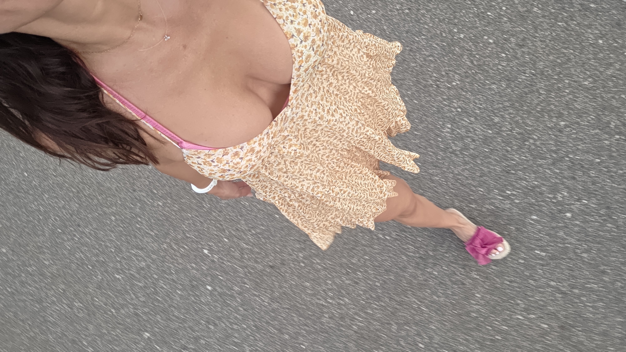 A woman is standing on an asphalt surface, possibly a road or path. She is wearing a short, floral-patterned dress with a pink strap. The dress is light yellow with small, darker floral prints. She has dark hair, and is wearing a necklace and a white bracelet. On her left foot, she is wearing a pink slip-on sandal with a fluffy bow. The image is taken from a high angle, looking down at the woman. The background is a blurred asphalt surface, indicating motion or a shallow depth of field.
