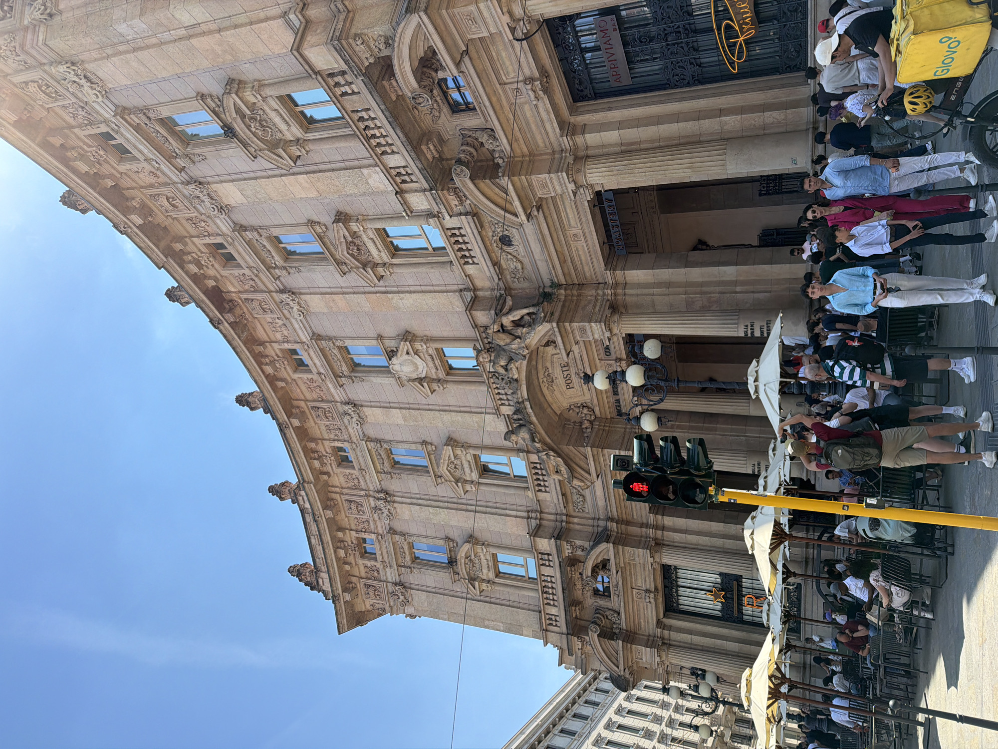 Here is a description of the image:
The photograph captures a bustling street scene in Milan, Italy, likely near a landmark building. The building, which takes up a significant portion of the frame on the left, features classical architecture with ornate details, windows, and columns. "POSTE" is emblazoned above the entrance. A coffee roastery is adjacent to the "Poste".
People are walking on the sidewalk and seated at tables under white umbrellas, which suggests an outdoor café setting. A yellow Glovo delivery bag is visible, indicating the presence of food delivery services in the area. The traffic lights are illuminated red. A line of people is waiting to enter the "Poste" building.
The sky is blue, indicating it is a sunny day. Other buildings are visible in the distance, offering depth to the urban landscape.