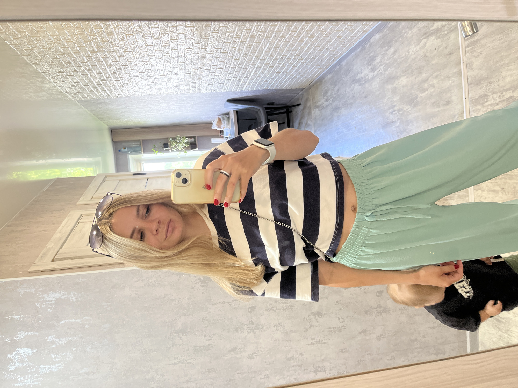 A blonde woman with sunglasses perched on her head and wearing a striped t-shirt and light green pants is taking a selfie in a mirror. Her right hand, painted with red nail polish, is holding a smartphone. A toddler in a black t-shirt is partially visible in the reflection behind her. The environment appears to be an indoor space, possibly a hallway or a room with a mirrored wall, featuring textured wallpaper and a brick-patterned wall in the background.